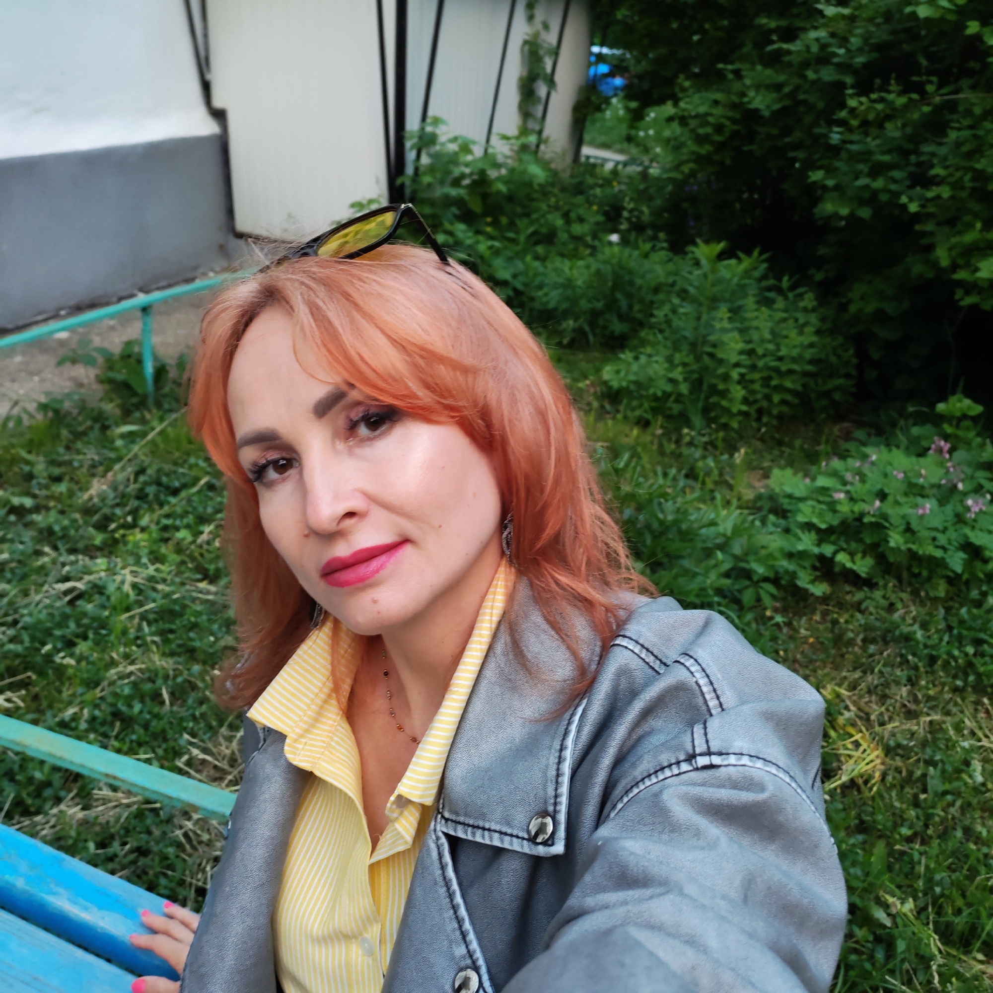 A woman with red hair and fair skin sits on a blue bench surrounded by green foliage. She is wearing a yellow striped shirt and a gray jacket. Sunglasses are perched on top of her head. The setting appears to be outdoors near a building. She is located in the city of Chaikovsky, Russia.