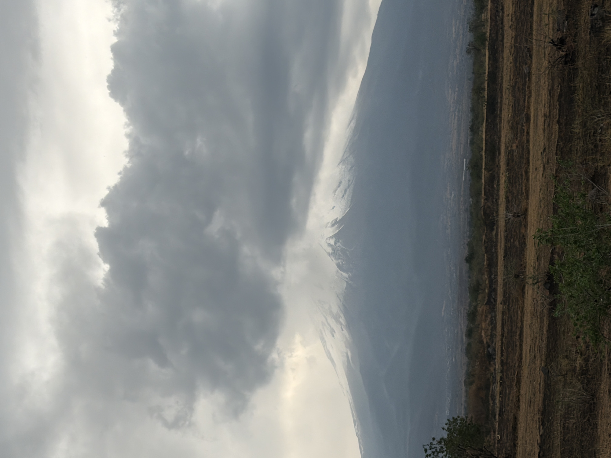 The image depicts a mountain obscured by heavy clouds, with evidence of snow or ice near the peak. The foreground shows a landscape with terraced fields, appearing dry or dormant. Vegetation is sparse, mainly consisting of grasses and small bushes. The location is identifiable as Metepec, Mexico, due to the distinctive landscape features typical of the region. The overall impression is one of a landscape shrouded in overcast conditions.