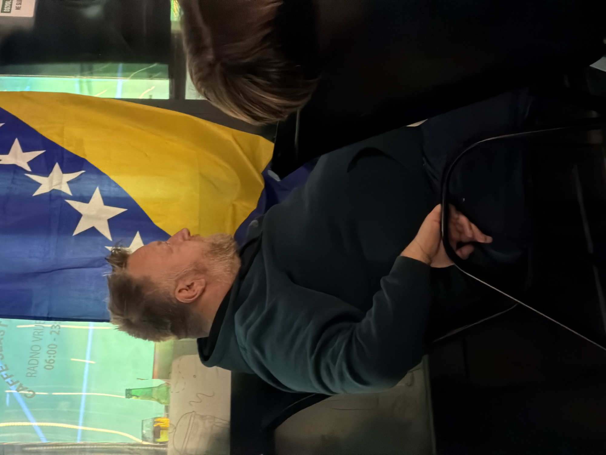 In Nević Polje, Bosnia and Herzegovina, a middle-aged man with a beard is seated in a chair, facing right. He is wearing a dark teal hooded sweatshirt and dark pants. His arms are resting on his lap, with his left hand partially visible. To his right, a Bosnian flag is displayed prominently. In the background, a window shows a green-tinted advertisement for "CAFFE SLAS." with operating hours "RADNO VRIJEME 06:00-23:00." A glass with a yellow liquid and a green bottle are also visible on a surface behind the man. The scene appears to be indoors, possibly in a cafe or public establishment, with other patrons hinted at by the presence of another person's head and shoulders to the right of the frame.
