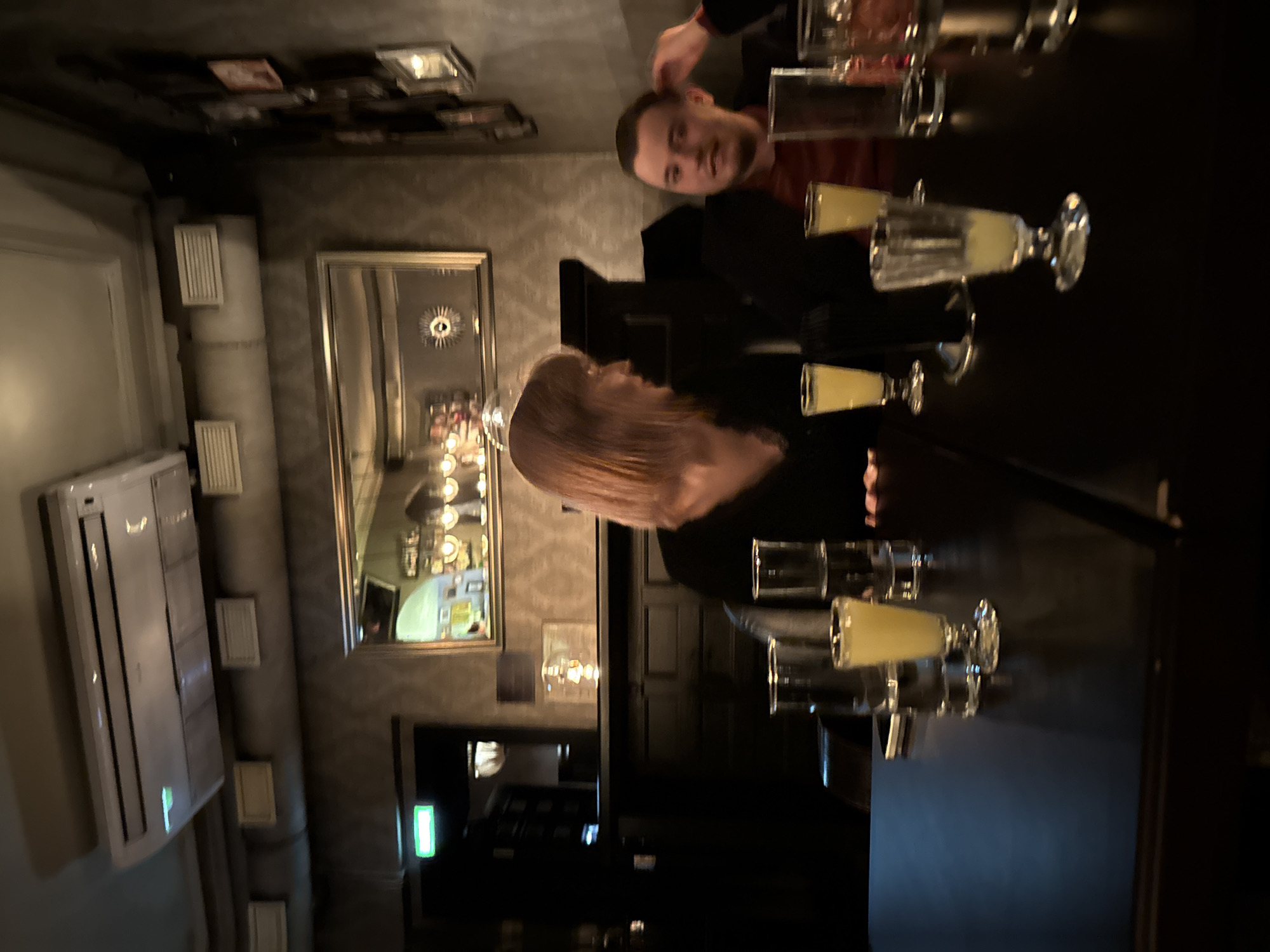 The image captures an indoor scene, possibly in a restaurant or bar. Two individuals are present at a table; one is male with a beard and short hair, while the other is female with shoulder-length blonde hair. Both are partially visible in the frame.
The table is set with multiple glasses, some containing a yellowish liquid, suggesting they might be alcoholic beverages. The dark surface of the table reflects light, adding to the dimly lit ambiance.
The background reveals a wall with a decorative pattern and a large rectangular mirror reflecting the interior, including what appears to be a chandelier or ornate lighting fixture. An air conditioning unit is mounted on the wall to the left.
Due to limited visual information, the exact location within Moscow, Russia, cannot be determined.