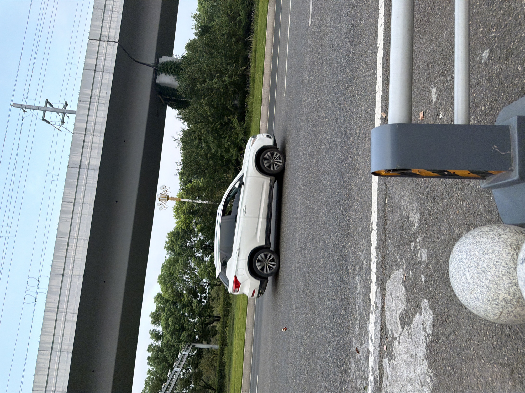 A white SUV drives on a paved road under a large overpass. The overpass is supported by concrete pillars, and overhead power lines are visible. Trees and greenery line the side of the road. In the foreground, a portion of a gray metal guardrail with yellow and black reflective tape is visible, along with a rounded gray stone bollard. The road surface is asphalt with white lane markings and some patchy discoloration. A small, reddish-brown object, possibly litter, is on the road to the left of the SUV. The sky is overcast. The location is Jiangjin, China.