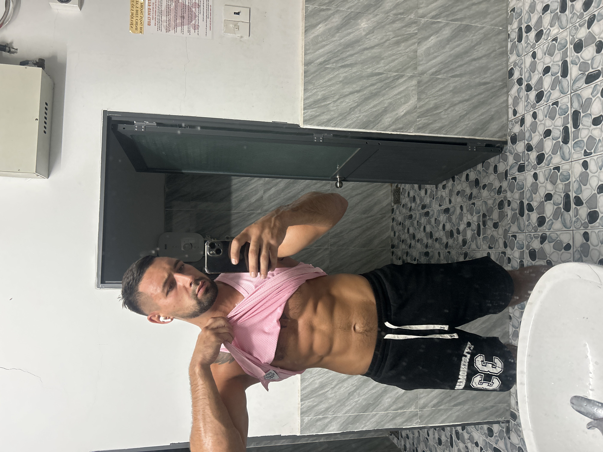 A man stands in front of a mirror, lifting his pink tank top to reveal his abdominal muscles. He is wearing black shorts with white trim and the number "33" on the left leg. The background shows a tiled wall and floor, a closed door, and a sink in the foreground. A notice is posted on the wall to the right of the door. The location is identified as Nha Trang Ward, Vietnam.