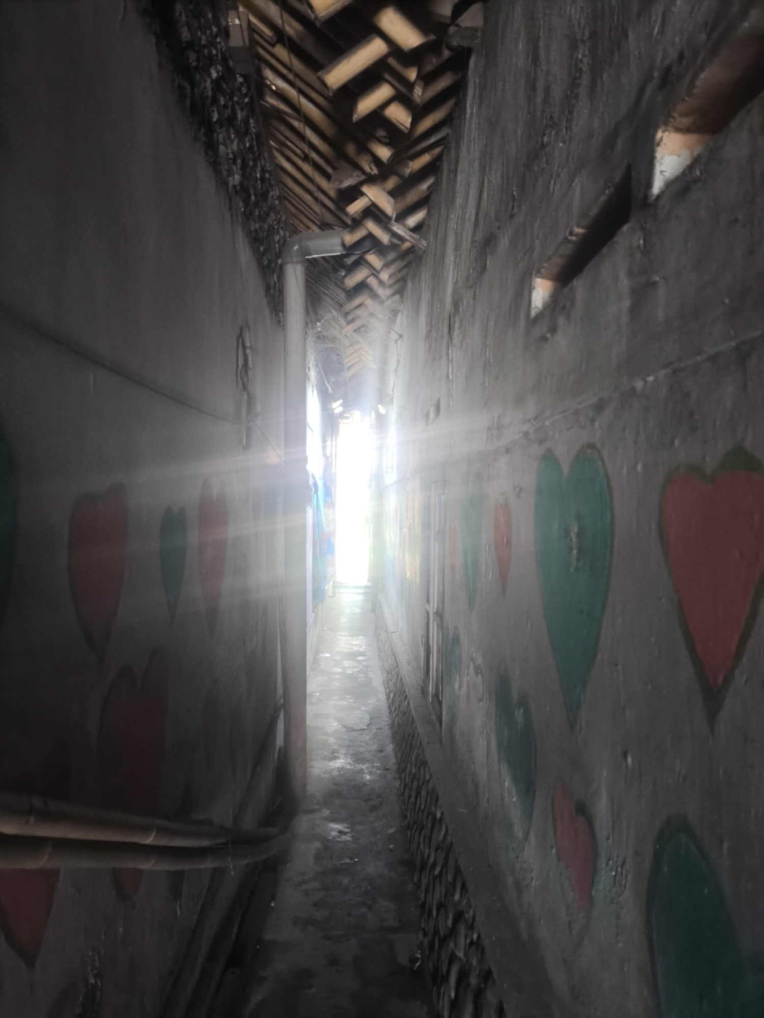 The image depicts a narrow alleyway in Lumajang, Indonesia. The walls on both sides of the alley are textured and appear to be made of concrete or stucco. The wall on the left has several heart shapes painted on it in red and green. The wall on the right also features painted heart shapes, predominantly in green and red. The alleyway leads to a brightly lit opening at the end, suggesting an exit or a more open area. The ceiling of the alley is constructed with wooden beams arranged in a herringbone pattern. There are no people visible in the image. The ground of the alley is paved and appears somewhat damp or wet. Several electrical conduits or pipes are attached to the wall on the left. The overall impression is of a somewhat weathered, possibly residential or community alleyway decorated with simple art.