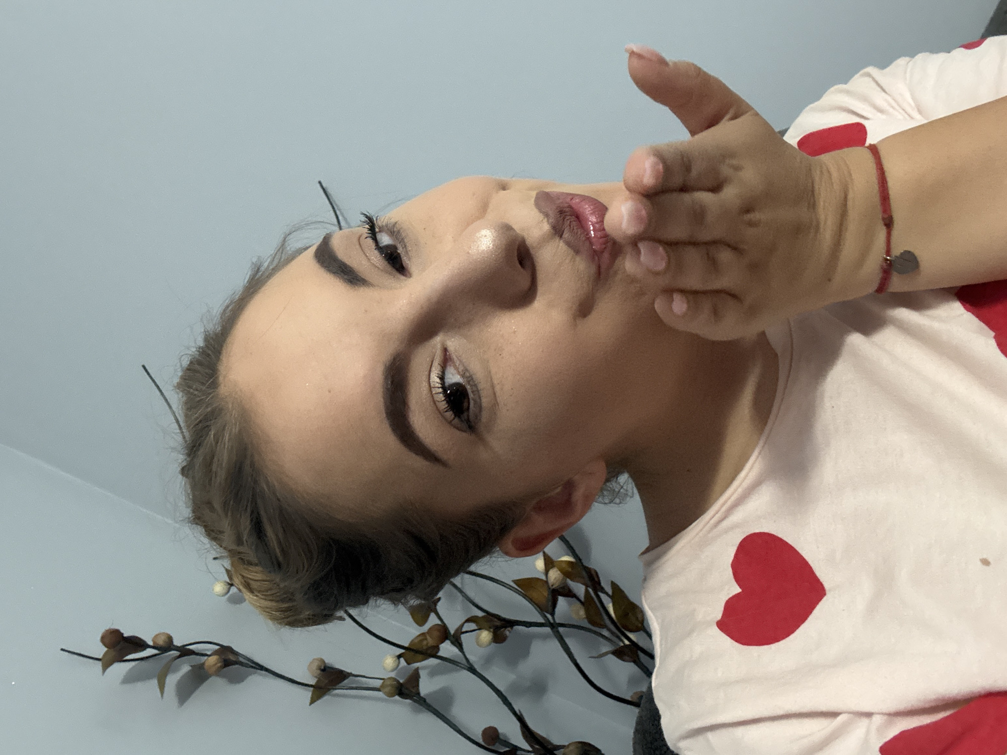 A medium close-up shows a light-skinned adult female, positioned slightly off-center to the right, in the city of Sofia, Bulgaria. She has light brown hair styled in an updo and is looking directly at the camera with a playful expression, her left eye slightly narrowed. Her lips are puckered, and her right hand is raised towards her mouth, fingers gently curled, in the gesture of blowing a kiss. She wears a light pink long-sleeved shirt adorned with red heart patterns, with one prominent red heart visible on her upper left chest. A thin red string bracelet with a small, metallic heart-shaped charm is present on her right wrist. Makeup includes defined eyebrows, mascara, and a light-colored lip gloss. The background consists of a plain light blue wall. To the left of the frame, behind her, a decorative artificial plant with dark branches, light brown berries, and muted green leaves is visible, casting a faint shadow on the wall.