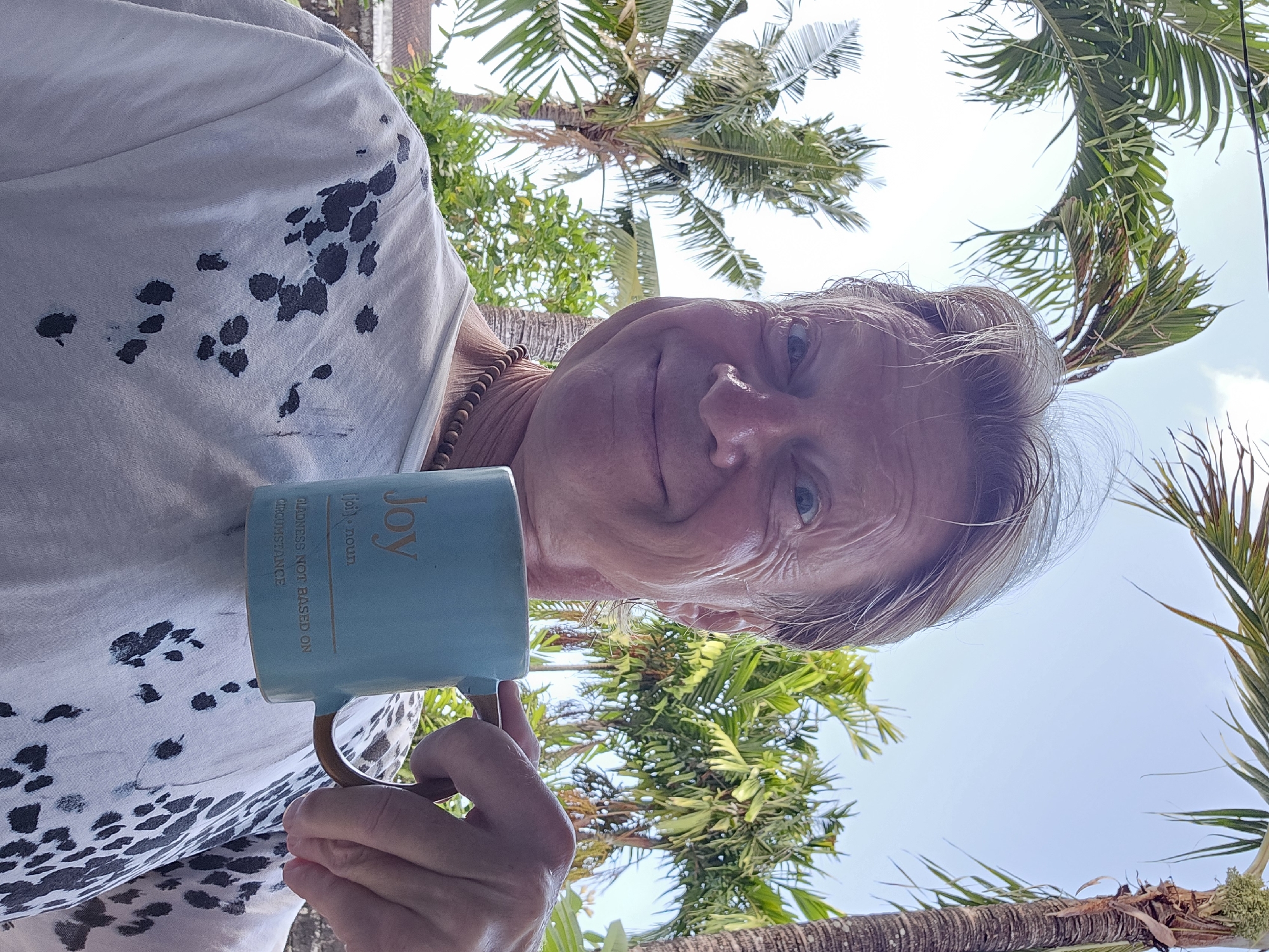 A man stands outdoors, holding a light blue mug with the word "Joy" and its definition printed on it. The man has blonde hair and is smiling broadly at the camera. He is wearing a white t-shirt with a black speckled pattern. He has a beaded necklace around his neck. The background shows lush green palm trees and a bright blue sky. The scene appears to be in Denpasar, Indonesia, given the presence of tropical vegetation common in that region.