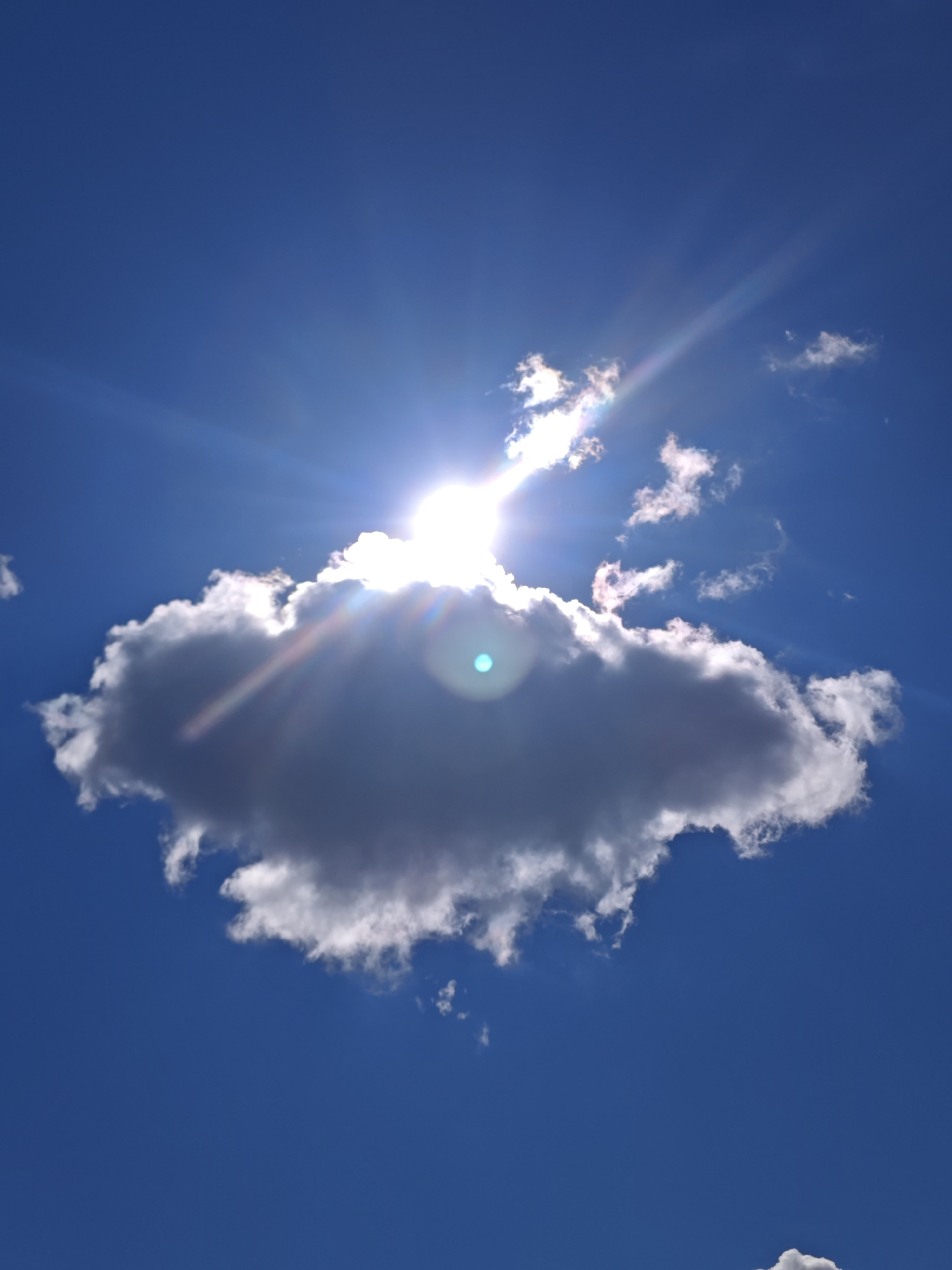 A low-angle, vertical shot captures a bright sun partially obscured by a large, dark cloud in a clear blue sky. Sunbeams radiate outwards from behind the cloud, creating lens flare. Several smaller, wispy clouds are scattered throughout the sky. The scene is devoid of any human presence or recognizable urban elements, making it impossible to determine its location within Moscow, Russia, or any other city.