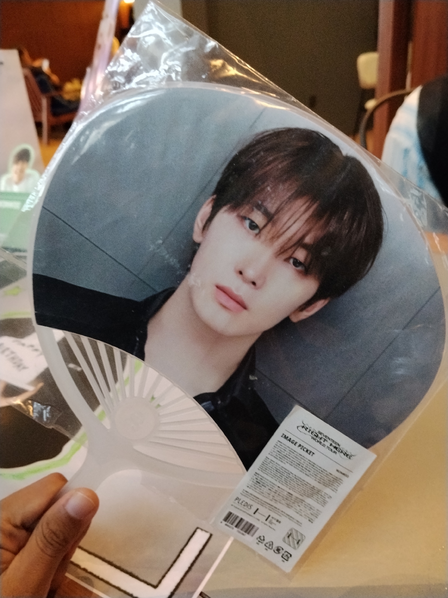 A hand is holding a round fan with an image of a young East Asian male printed on it. The fan is inside a clear plastic packaging. The male in the image has dark hair with bangs and is wearing a black shirt. A white label with text and a barcode is attached to the plastic packaging near the bottom right of the fan. The background is blurred and appears to be an indoor setting with warm lighting, possibly a retail store or event venue. There is no information in the image that indicates the location in the city of Serang, Indonesia.