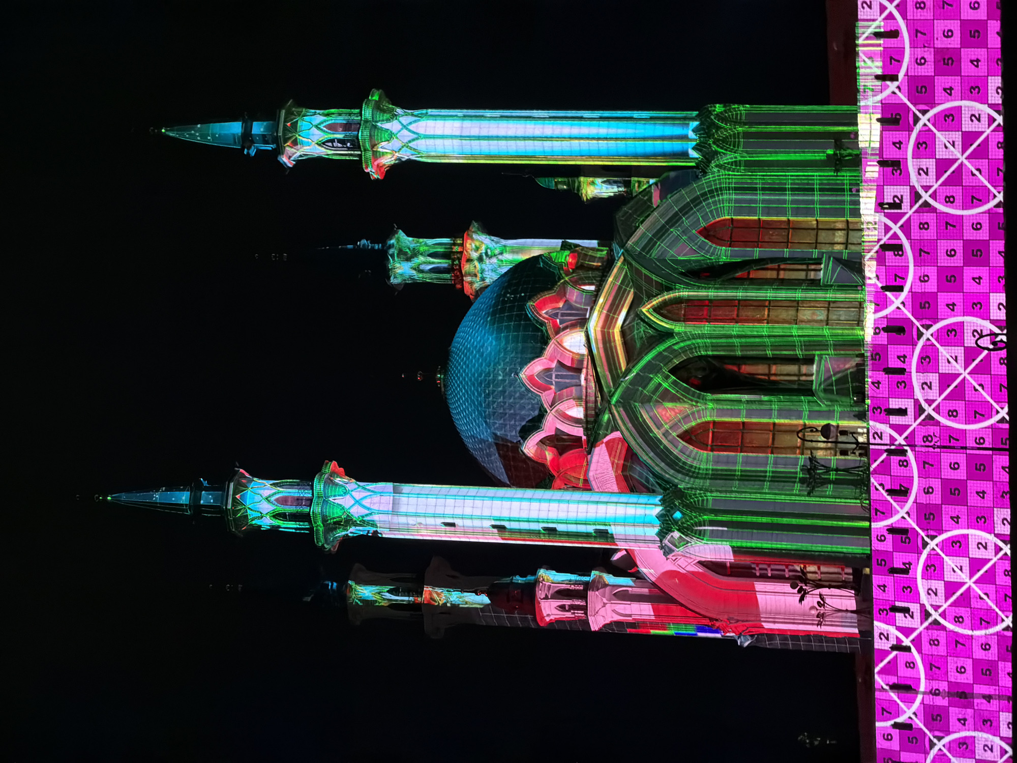 Here is a description of the image:
The image showcases a nighttime view of a mosque-like structure with a central dome and two minarets. The building is illuminated with a vibrant and complex light display, featuring a grid pattern of green lines, red highlights on the dome, and blue accents on the minarets. The right side of the image is dominated by a bright purple section with a geometric pattern and numbers. The sky is dark, emphasizing the brightness and colorfulness of the structure and its projections. Based on the architecture and color schemes, this is the Qolsharif Mosque in Kazan, Russia during a light show. The image is captured from a low angle, adding to the building's imposing presence. The composition is somewhat unconventional, with the structure rotated and elements like the purple section on the right dominating the frame.