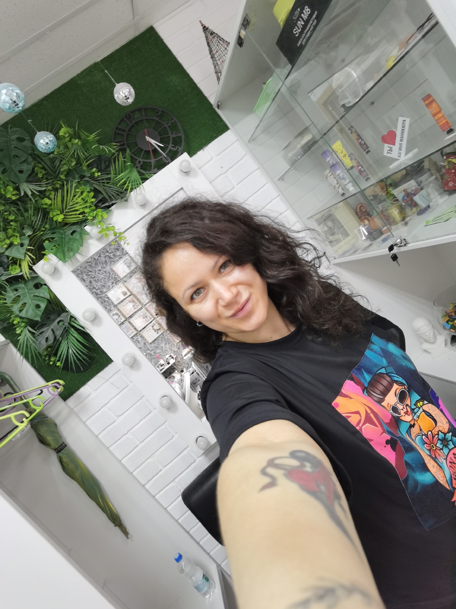 The image shows a selfie taken by a woman with curly dark hair and a tattoo on her arm. She is wearing a black t-shirt with a colorful graphic print. The background includes a wall with white brick texture, artificial plants, a clock, disco balls, a mirror with lights, and a glass display cabinet filled with various items. A green umbrella and a bottle of water are visible on a shelf. The scene appears to be indoors, possibly in a beauty salon or a similar establishment, given the mirror with lights and the display cabinet. The specific location in Chaikovsky, Russia cannot be determined from the image alone.