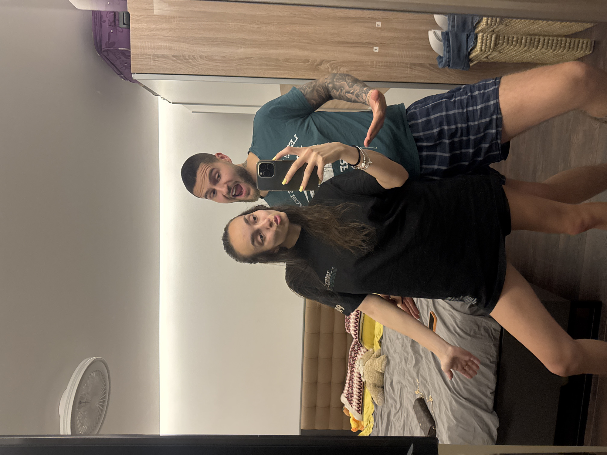 A mirror selfie, taken by a man and a woman, captures their reflections in what appears to be a bedroom in Sofia, Bulgaria. The image is oriented with the top of the mirror to the left. The woman, with long dark hair, holds a dark-cased smartphone to take the picture, her fingernails painted yellow. She wears a black t-shirt with illegible mirrored text and dark shorts, appearing to make a slight pout.
Behind her, a man with short dark hair and a beard makes a playful open-mouthed expression. He wears a dark teal t-shirt, which has the word "LED" and other mirrored text visible, and blue plaid shorts. His left arm is extensively tattooed.
The background shows a modern bedroom setting. A wooden wardrobe or cabinet with white accents is prominent. On top of the wardrobe, a purple suitcase-like item is visible, along with woven baskets containing what appear to be shoes. A small white fan rests on a surface to the left of the frame. To the right and below, a bed with a brown padded headboard, grey bedding, a patterned blanket, and a white stuffed animal are visible. The overall mood is casual and playful.
