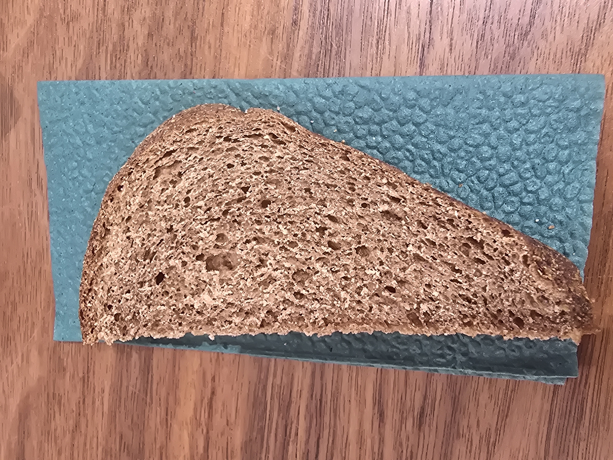 A slice of whole wheat bread lies on a teal-colored, textured surface, which in turn rests on a wooden table. The bread is positioned diagonally, with its crust facing the lower left corner and its soft interior facing upwards. The wooden table has a prominent grain pattern, running horizontally across the image. The scene is lit from above, casting subtle shadows. There is no indication of the location in Ulan-Ude, Russia.