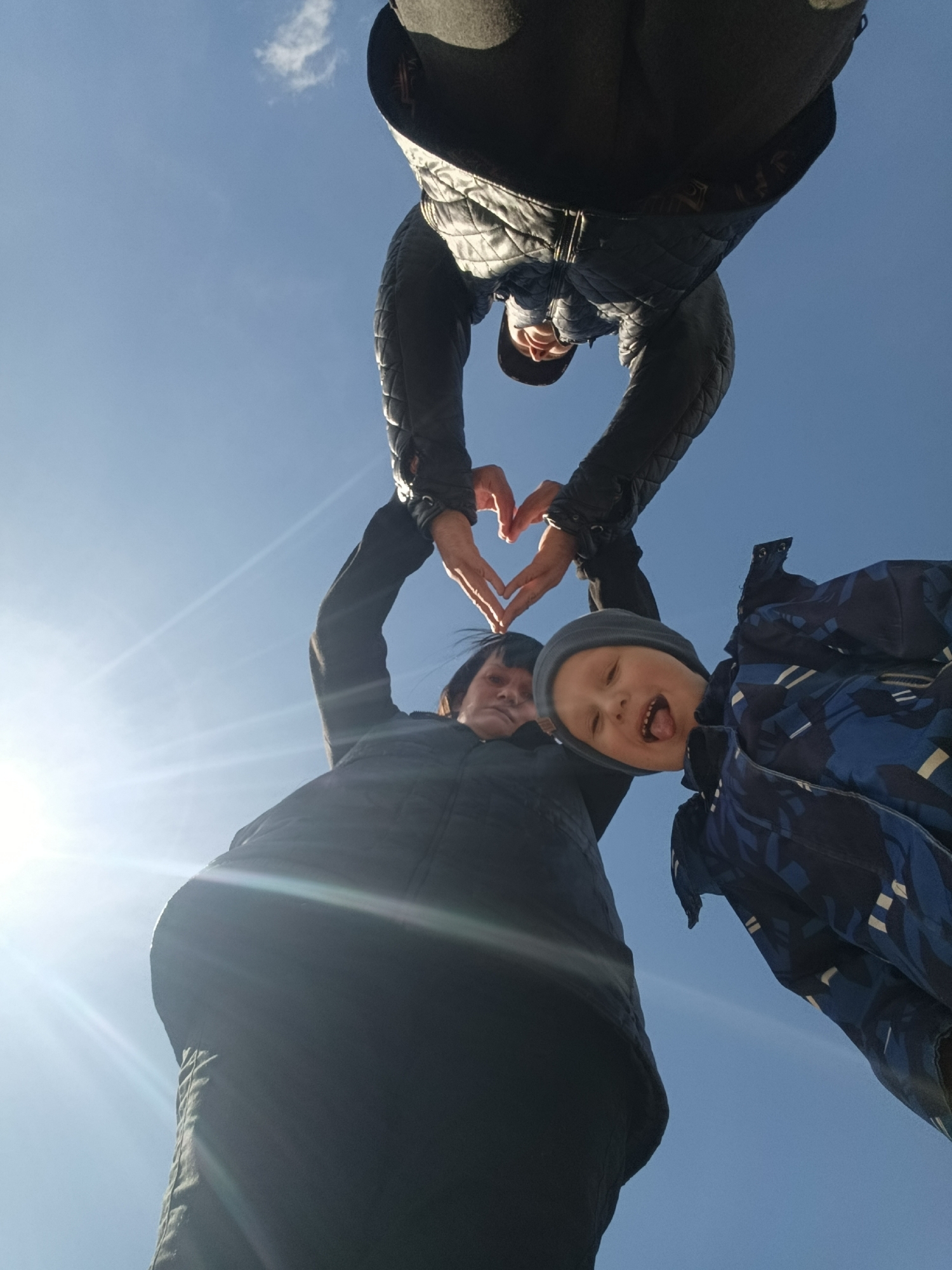 A low-angle shot shows three individuals against a clear blue sky. The topmost individual wears a dark quilted jacket, their hands forming the top of a heart shape. Below them, another person, possibly a woman, wearing a black puffer coat with their arms raised so their hands connect with the top individuals hands forming the heart shape. To the right, a young child wearing a blue patterned jacket and a gray knit hat sticks their tongue out. The photo appears to have been taken outdoors on a sunny day. The city location provided seems incongruous with the scene depicted.