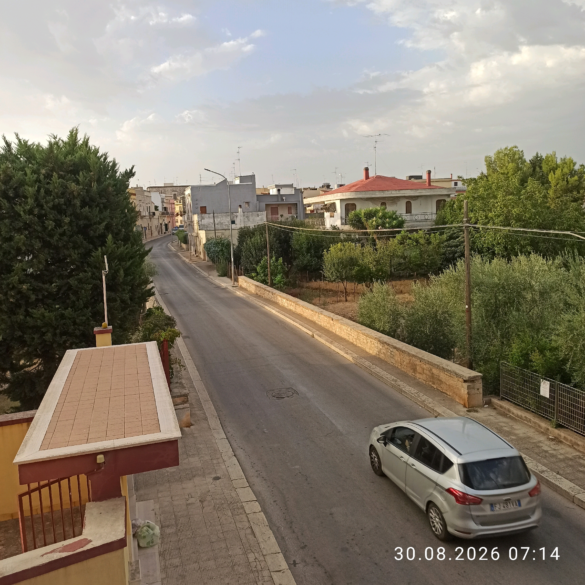 The image shows a street scene in Bitonto, Italy, on August 30, 2026, at 07:14. A silver minivan is traveling down the road, and there is a building with a red-tiled roof in the background. The street is lined with trees and houses, and there is a stone wall alongside the road. The sky is cloudy. The scene appears to be a residential area.