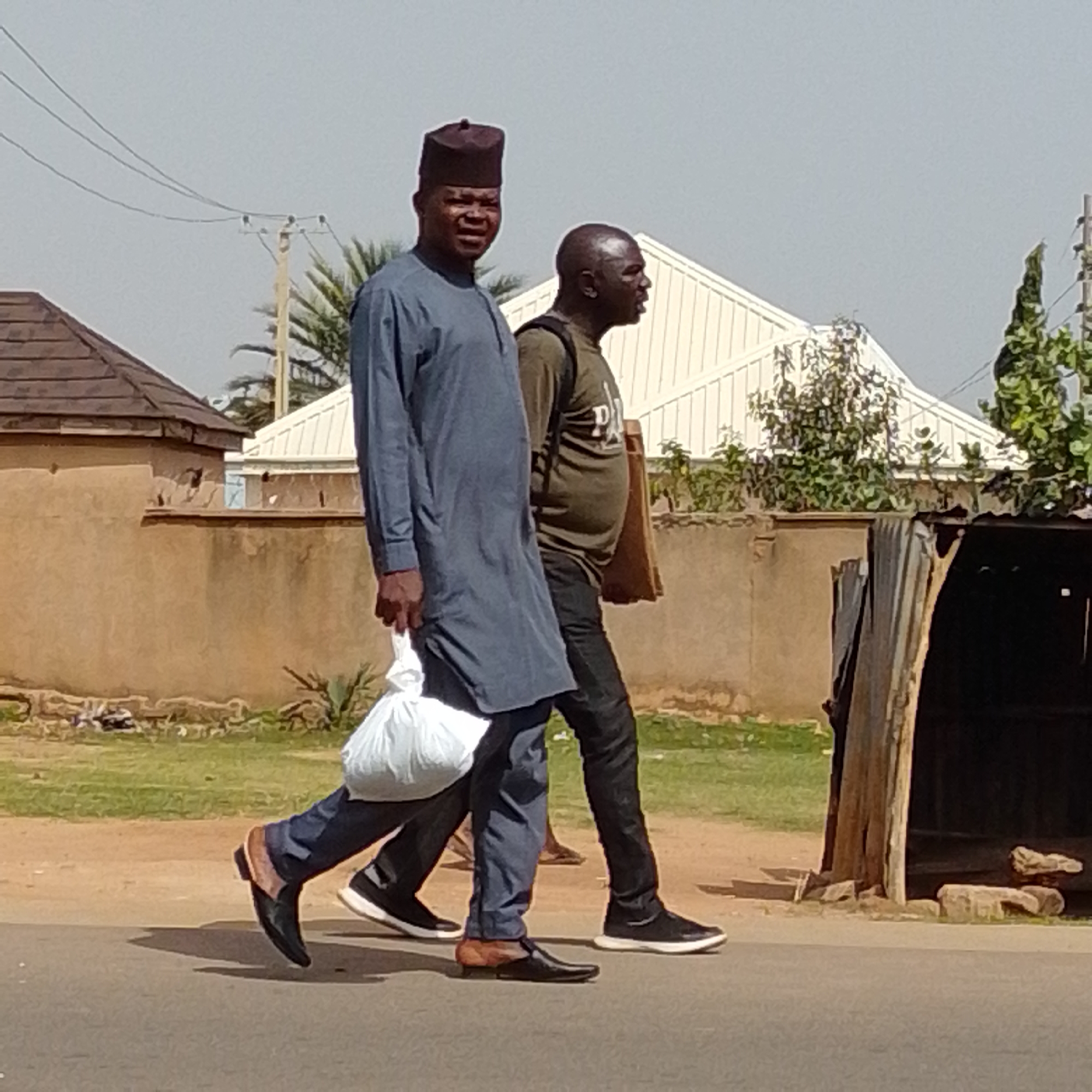 Two men are walking on a paved road in Tempure, Nigeria. The man in the foreground is wearing a blue tunic and a dark, tall cap. He is carrying a white plastic bag in his left hand. He is wearing black dress shoes. The man behind him is wearing a green t-shirt and black pants. He has a dark bag or backpack on his shoulders. He is wearing black sneakers. The background shows a brown wall, some greenery, and buildings with white, ribbed roofs. The sky is clear and light blue.