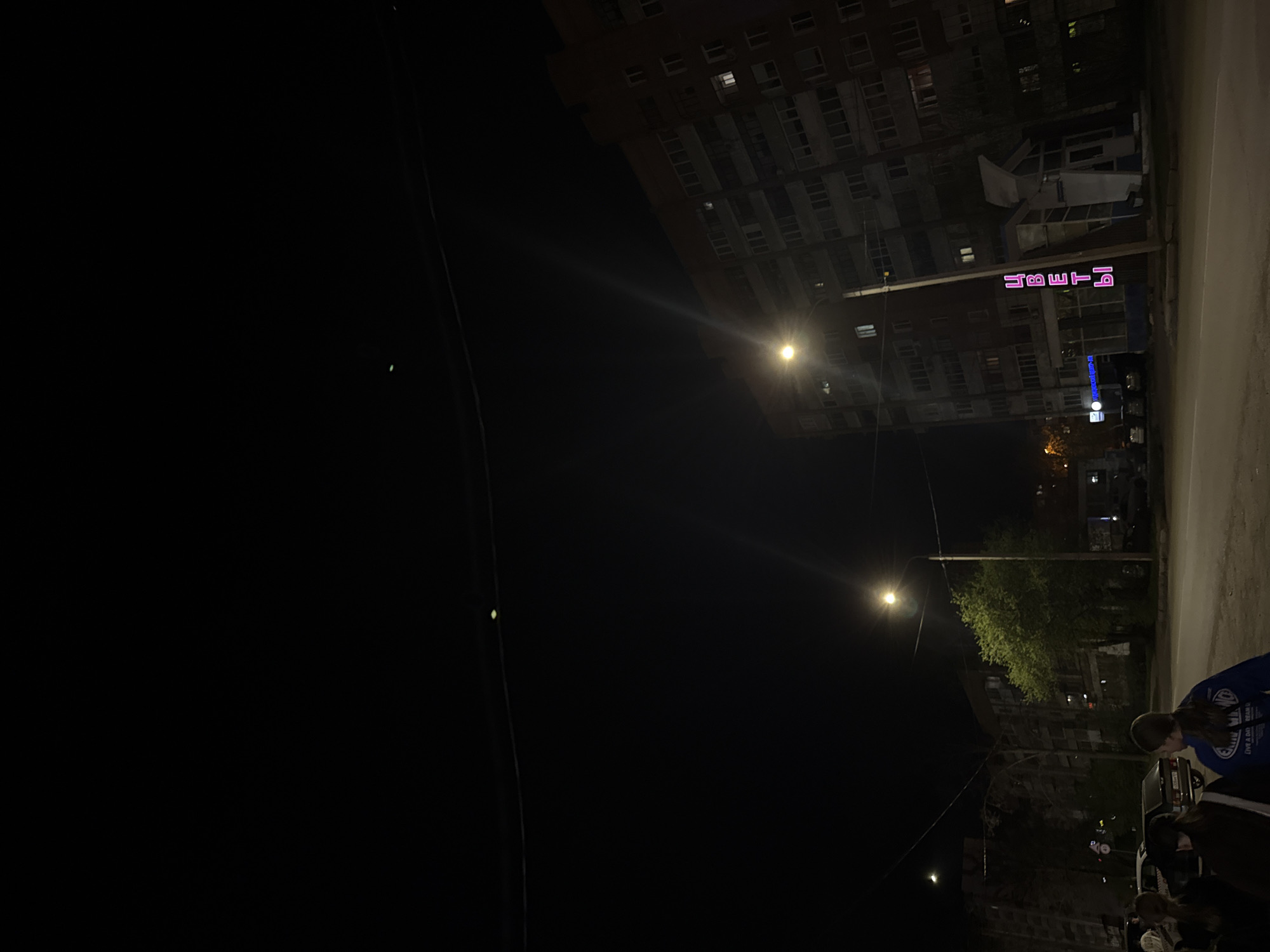 The image shows a nighttime scene in Perm, Russia. A tall apartment building is visible in the background, with a neon sign that reads "ЦВЕТЫ" (flowers). Street lights illuminate the scene. A street and sidewalk are also present. People are visible on the sidewalk near parked cars. Wires are visible in the dark upper left corner.