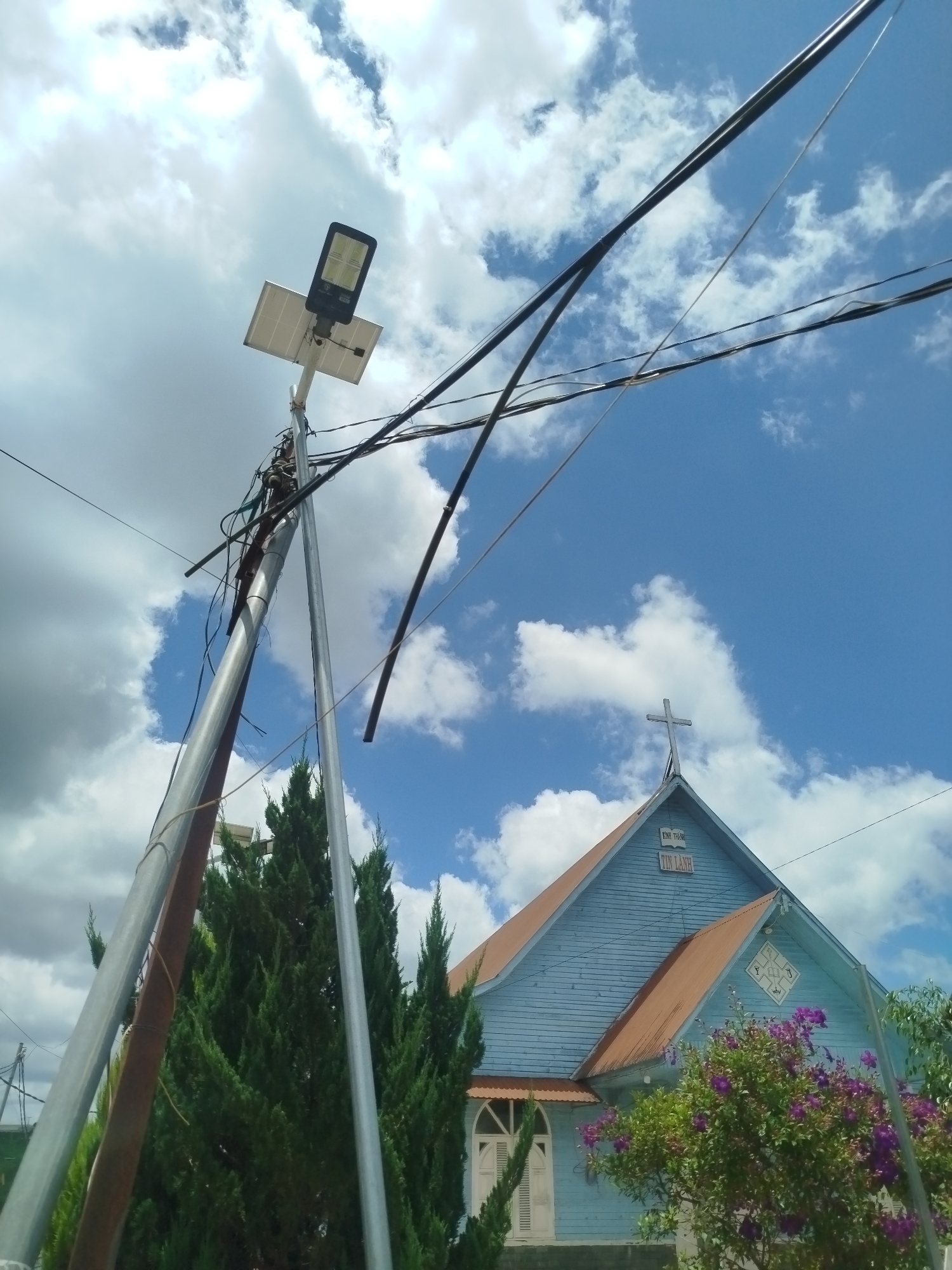 This is a photograph taken from a low angle, looking up at a bright blue sky with scattered white clouds. In the foreground, a tall metal pole supporting a solar-powered street light and a tangle of electrical wires dominates the left side of the frame. The pole is silver and appears weathered, with several wires of varying thickness attached to it, some dangling precariously. Behind the pole, to the right, is a light blue wooden church with a copper-colored roof. A cross is visible on the peak of the roof. Text is present above the cross, in Vietnamese, with the words "TIN LANH" clearly legible. In front of the church, there are several evergreen trees and a bush with purple flowers. The overall impression is of an outdoor scene, likely in a rural or semi-rural area of Thôn 6, Vietnam, featuring both modern infrastructure and traditional architecture. There are no people visible in the image.