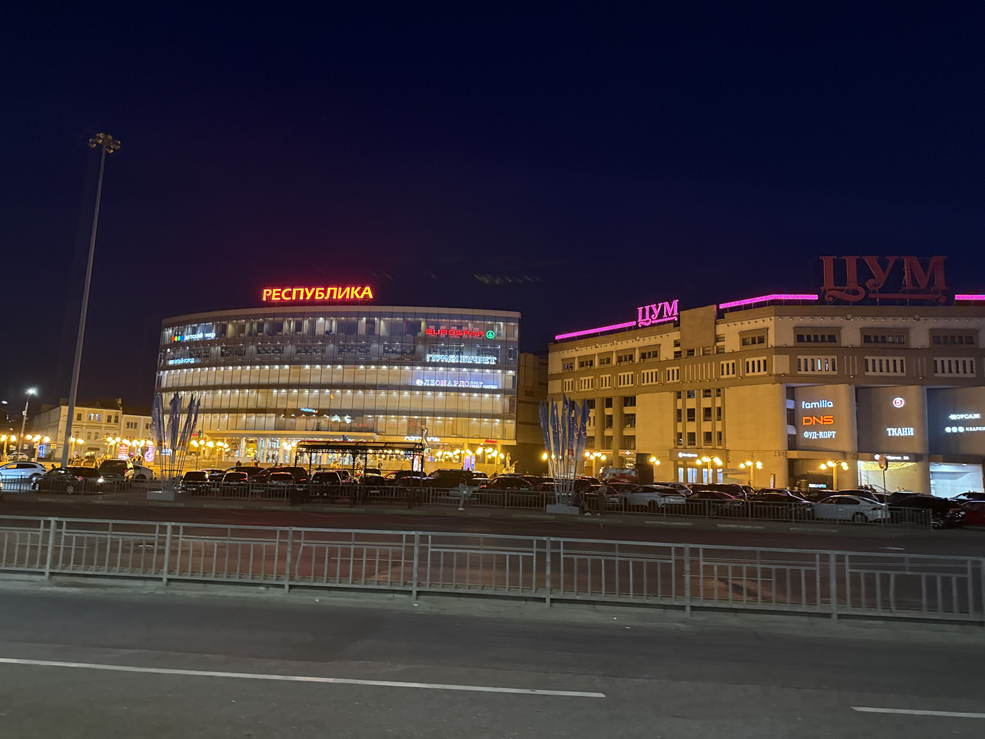 The image captures an urban street scene at dusk or night in Nizhny Novgorod, Russia. Prominently featured are two large, illuminated buildings displaying neon signs. The building on the left has a red neon sign that reads "РЕСПУБЛИКА" (Respublika), and below it, signs for "ДЕТСКИЙ МИР" (Detskiy Mir - Children's World), "EUROSPAR", and "ГУРМАН БУФЕТ" (Gourmet Buffet). This building is modern with a glass facade reflecting the lights. To its right is another building with a neon sign that says "ЦУМ" (TSUM - a department store) in pink and red lettering. This building appears to be older, with a more classical architectural style and rows of windows.
In front of the buildings, a street is visible with several cars parked and moving. A metal fence runs along the sidewalk, separating it from the street. Streetlights are on, casting a warm glow. The sky is a deep blue, indicating the late hour. The overall atmosphere is one of a busy commercial district at night.