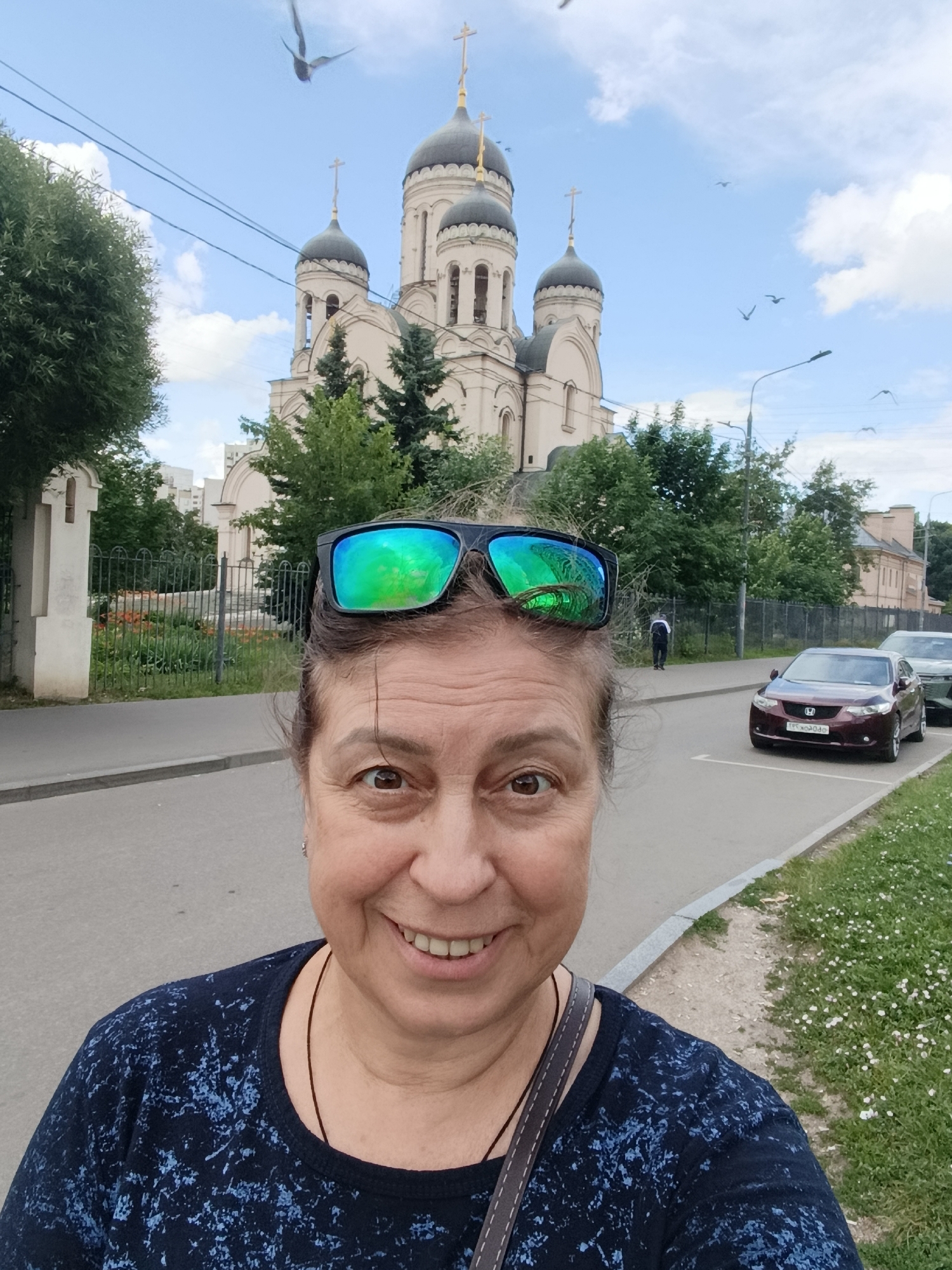 A middle-aged woman with sunglasses perched on her head smiles at the camera in a selfie. She is wearing a dark blue shirt with a light blue pattern. In the background, a white church with dark domes and crosses stands against a cloudy blue sky. Several birds are flying in the sky. The church appears to be St. Nicholas Naval Cathedral in Kronstadt, Russia, not Moscow. A street with parked cars is visible in front of the church. A sidewalk and a grassy area with small white flowers are also present. The scene depicts a bright, daytime outdoor setting.