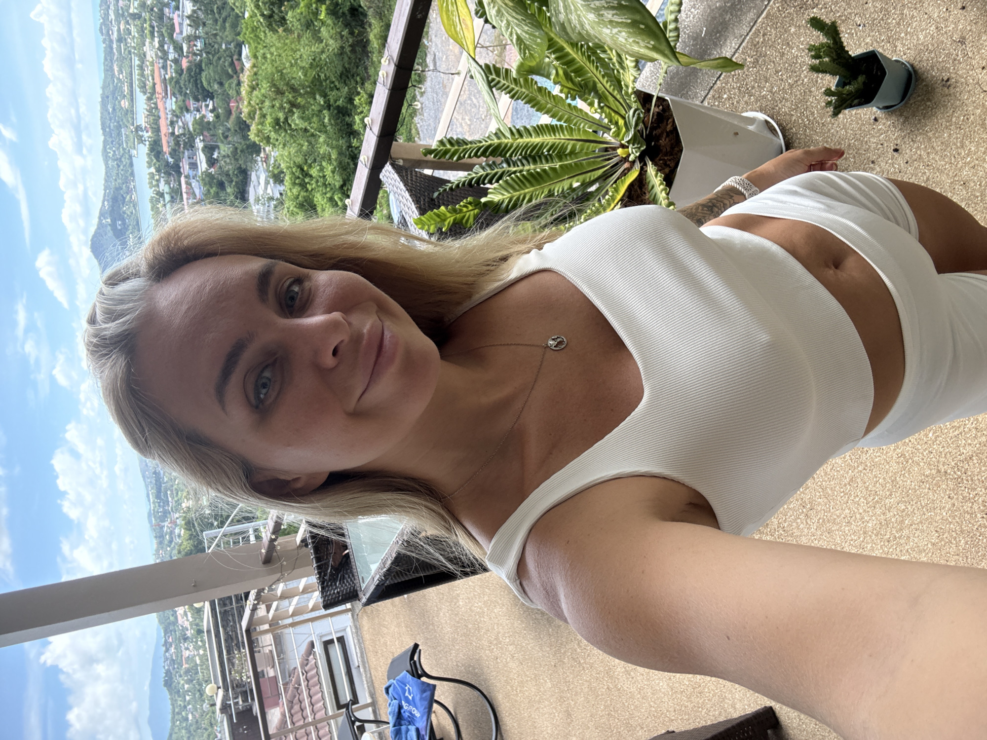 A blonde woman stands on a balcony overlooking a city with a bay and mountains in the distance, believed to be Baan Chaweng, Thailand. She is wearing a white crop top and white shorts and smiling at the camera. The balcony has a railing decorated with fairy lights, a table and chairs, a lounge chair with a blue towel, and several potted plants, including a large bird's nest fern and a small cactus. The sky is blue with scattered clouds.