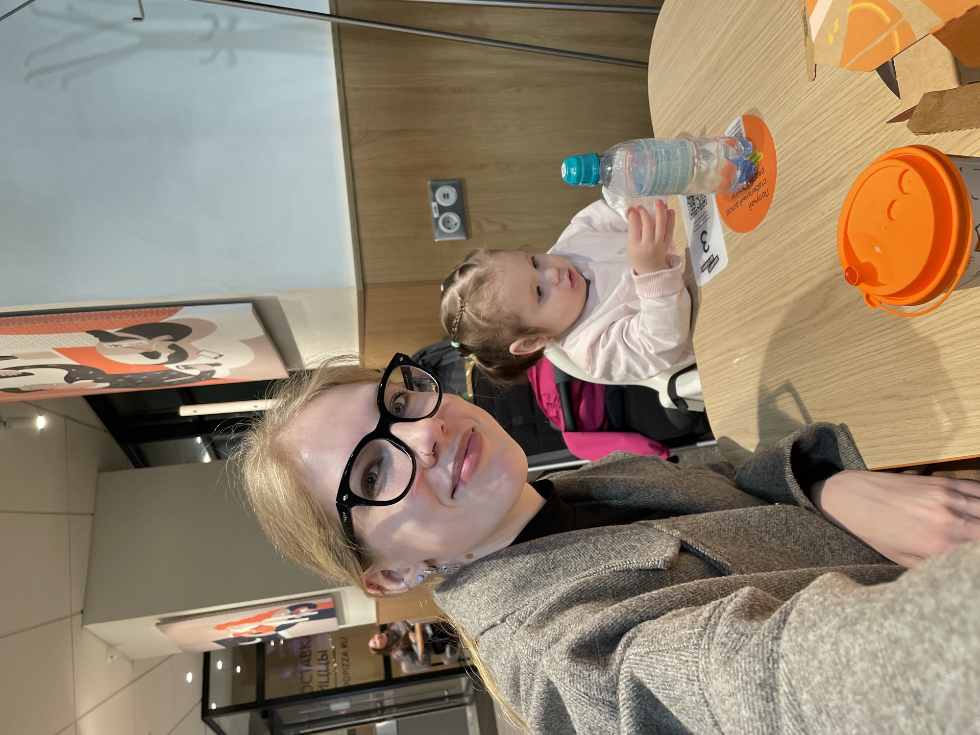 The image shows a woman taking a selfie with a young child at a table in a cafe or restaurant. The woman is wearing glasses and a gray jacket. The child is sitting in a high chair and reaching for a bottle of water. On the table are a fast food drink, a paper coaster, and other items. In the background, there are wall decorations, and the restaurant’s interior is visible through the windows. Based on the visible signage and the woman’s past social media posts, the location is identified as "Dodo Pizza" in Izhevsk, Russia.