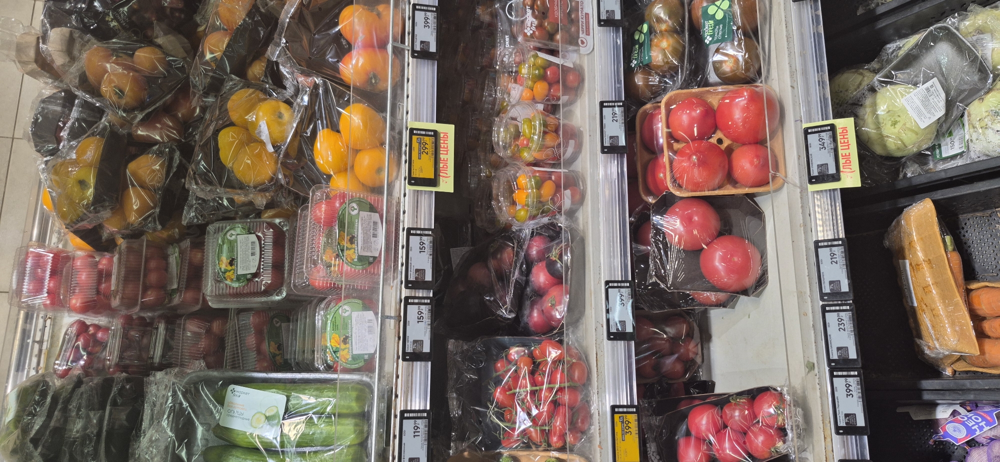 The image shows a refrigerated display case in a grocery store, likely in Saint Petersburg, Russia. The shelves are stocked with a variety of fresh produce, primarily tomatoes and cucumbers. There are many plastic containers and bags filled with different types of tomatoes, including cherry tomatoes, and several large red tomatoes. Cucumbers are also visible, packaged in clear plastic.
Price tags are attached to the front of the shelves, indicating that this is a retail environment. Some of the visible text on the price tags and packaging includes Russian words like "ОГУРЦЫ" (cucumbers), "ЧЕРРИ КУМАТО" (cherry kumato), and "УДОБНЫ" (convenient). There are also numbers that appear to be prices, such as "11999", "15999", "2199", "23999", and "34999". Some yellow price tags with the text "ЛЫЕ ЦЕНЫ" (discount prices) are also present.
The lighting suggests an indoor setting, typical of a supermarket, and the overall impression is one of abundance and freshness of produce. The image is taken from a slightly elevated angle, looking down into the display case. No people are visible in the frame.