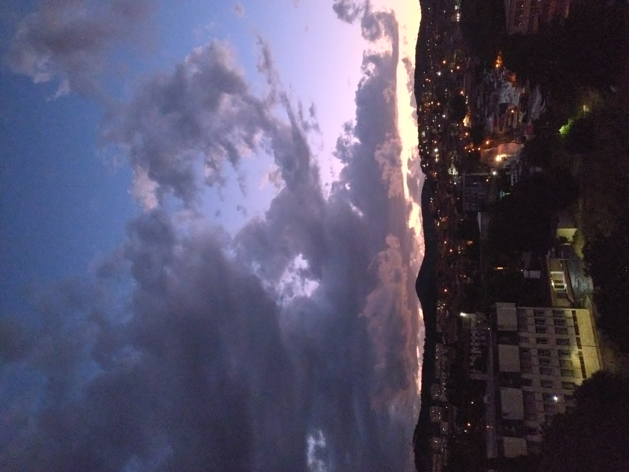 The image depicts a cityscape at dusk, likely Stara Zagora, Bulgaria, with a dramatic sky. The sky is filled with dark, heavy clouds transitioning to a soft lavender and light orange horizon. On the right side of the image, a hillside separates the sky from the city. A dense collection of buildings is visible, illuminated by streetlights and building lights. Some buildings are distinctly larger, possibly apartments or commercial structures. The lower right corner is obscured by trees and greenery, adding to the darkness in that part of the image. No people are visible.