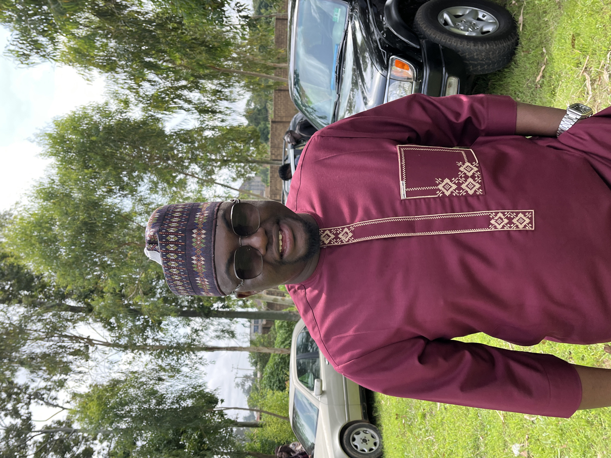 In Bukuru, Nigeria, a man stands outdoors in front of a group of trees and a dark SUV. He is wearing a maroon traditional outfit with a patterned cap and sunglasses. The man is smiling and appears to be posing for a photo. The background includes a light-colored sedan parked to the left of the SUV, and a few other people are visible in the distance. The ground is covered in green grass.