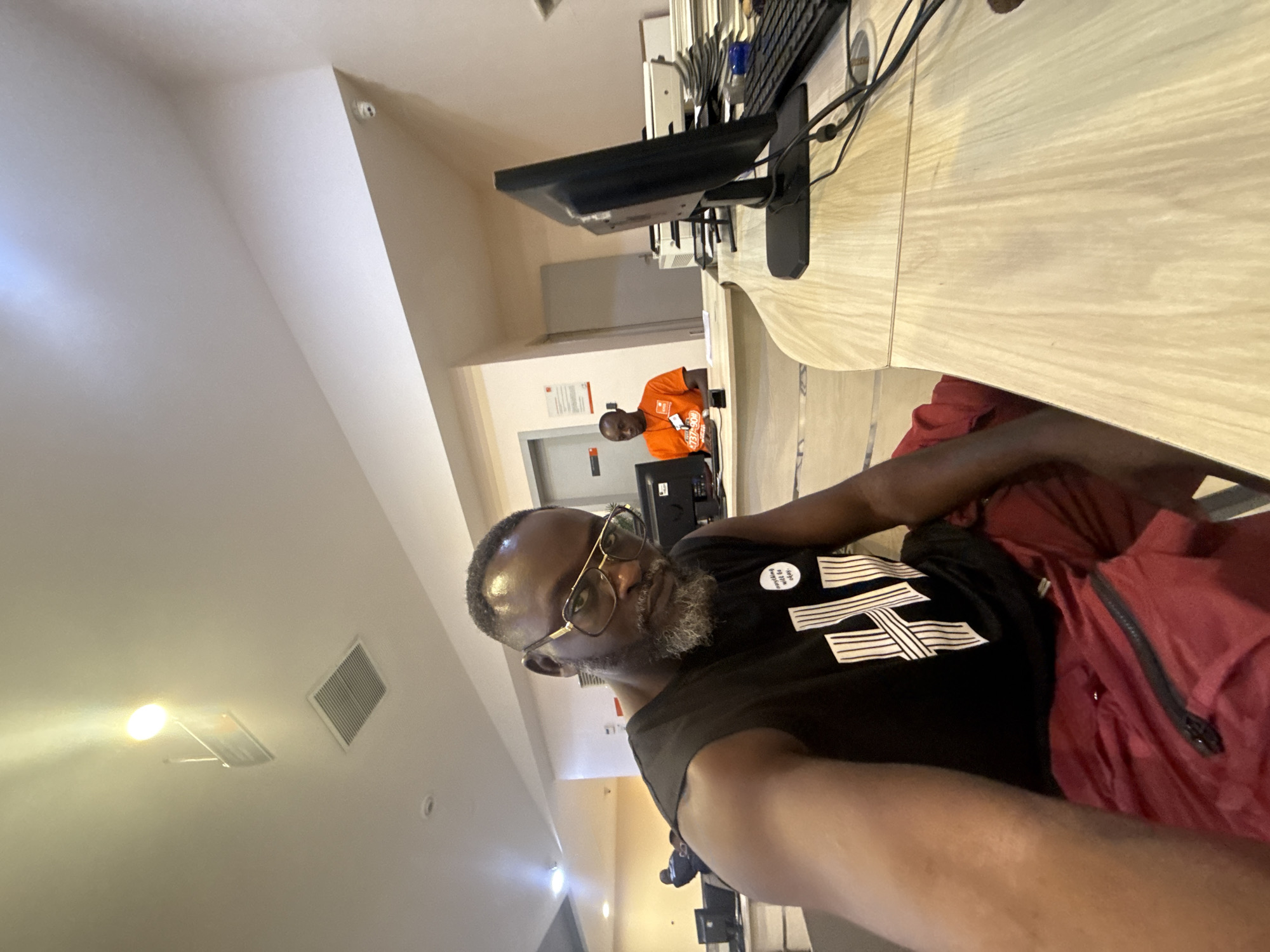 The image shows two men in what appears to be an office or computer lab setting. The foreground is dominated by a man wearing glasses, a black t-shirt with white horizontal lines, and red shorts. He has a graying beard and is looking at the camera. To his right, part of a red garment, possibly a jacket or bag, is visible.
In the background, another man is seated at a desk, wearing an orange t-shirt. He is looking down at a computer screen. The desk is made of light-colored wood and has computer equipment on it, including a monitor and keyboard. The room has a neutral-colored wall and a ceiling with light fixtures and a ventilation grate. One of the crops shows a security camera mounted on the ceiling. The lighting suggests it is daytime. The overall scene implies an indoor work or study environment. The text "love to be" is visible on a circular badge on the man's t-shirt.