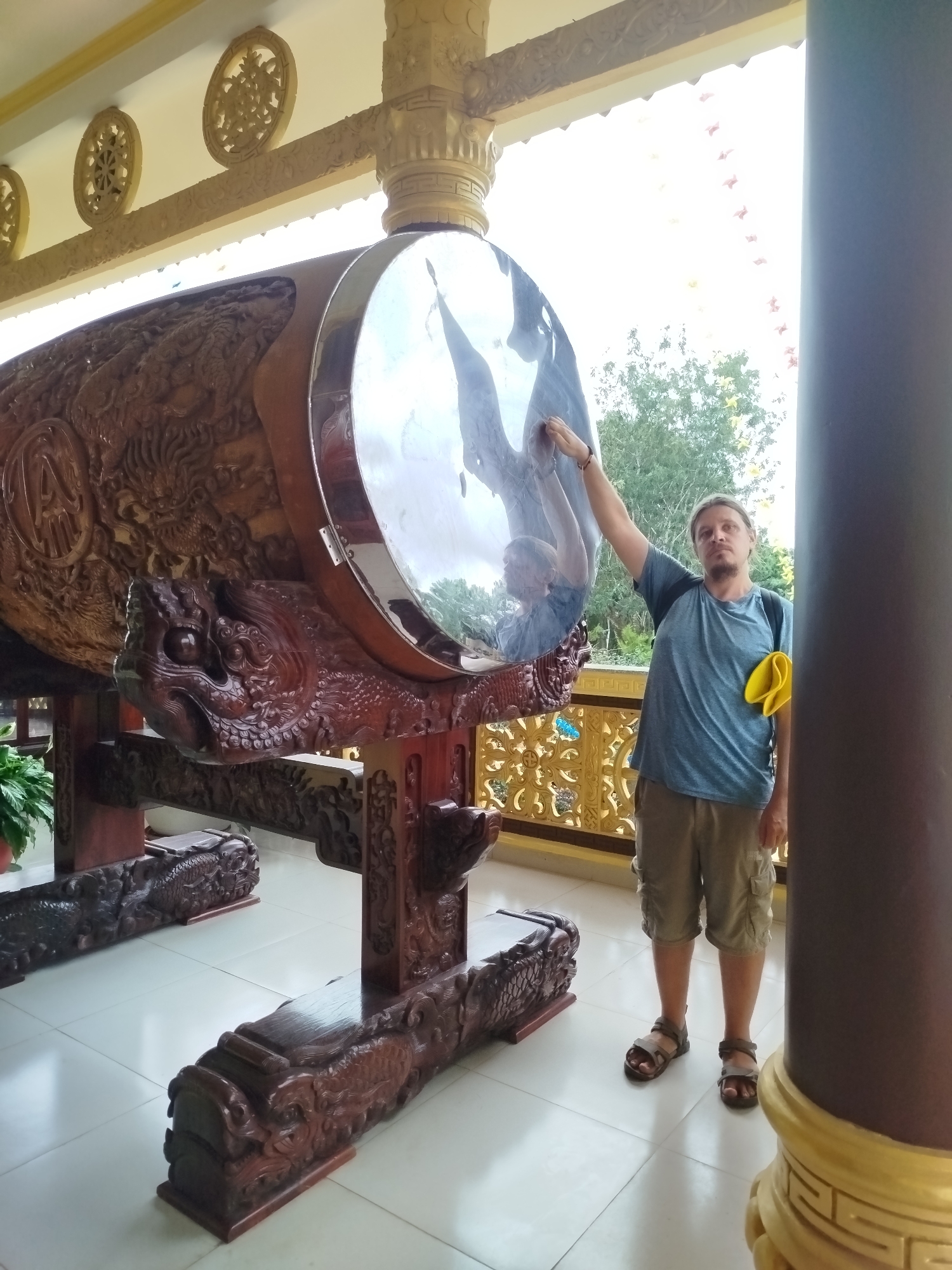 A man stands in a temple in Da Lat, Vietnam, posing with his reflection in a large, ornate drum. The drum is made of dark wood, carved with intricate designs of dragons and other mythical creatures. The man is wearing a blue t-shirt, khaki shorts, and sandals. He has a yellow cloth tucked into his waistband. The temple's architecture is visible in the background, with decorative carvings and columns. The floor is tiled in a light color.