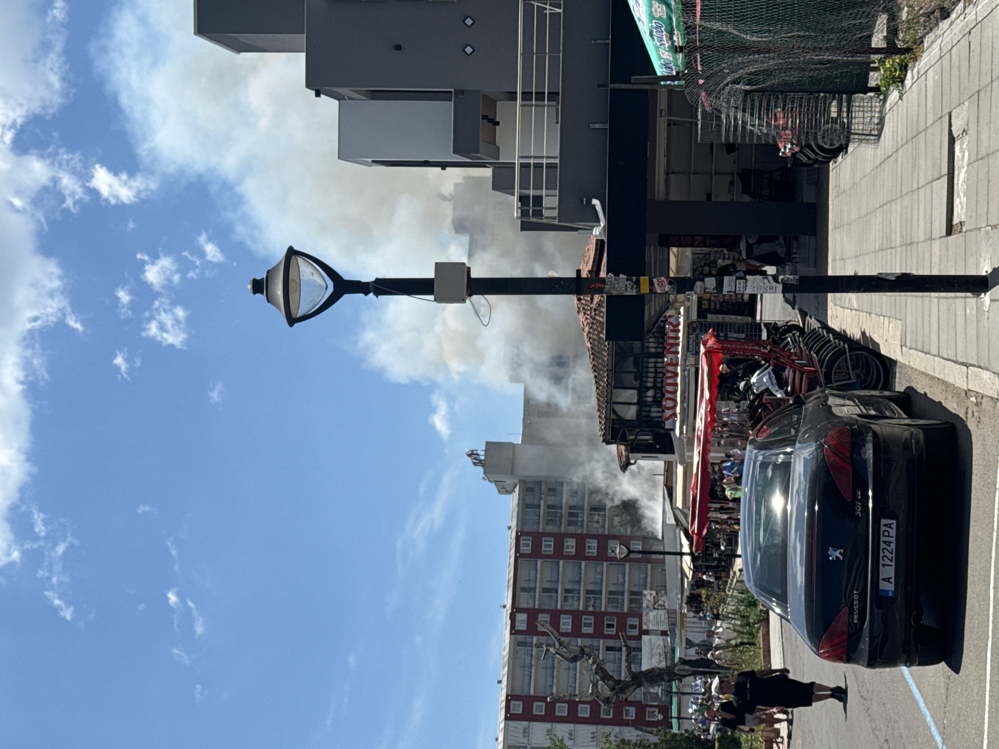 The image depicts a street scene in Nesebar, Bulgaria, likely during daytime. Smoke is billowing from a multi-story building in the background, indicating a fire or an emergency situation. In the foreground, a black Peugeot sedan is parked on the street. The car's license plate reads "A 1224 PA". A sidewalk lines the street, with a lamppost visible in the middle ground. People are walking on the sidewalk and in the street, some appearing to be onlookers. A store with a red awning is also visible, and bicycles are parked in front of another establishment. The sky is blue with scattered white clouds. The overall atmosphere appears to be one of concern due to the visible smoke.