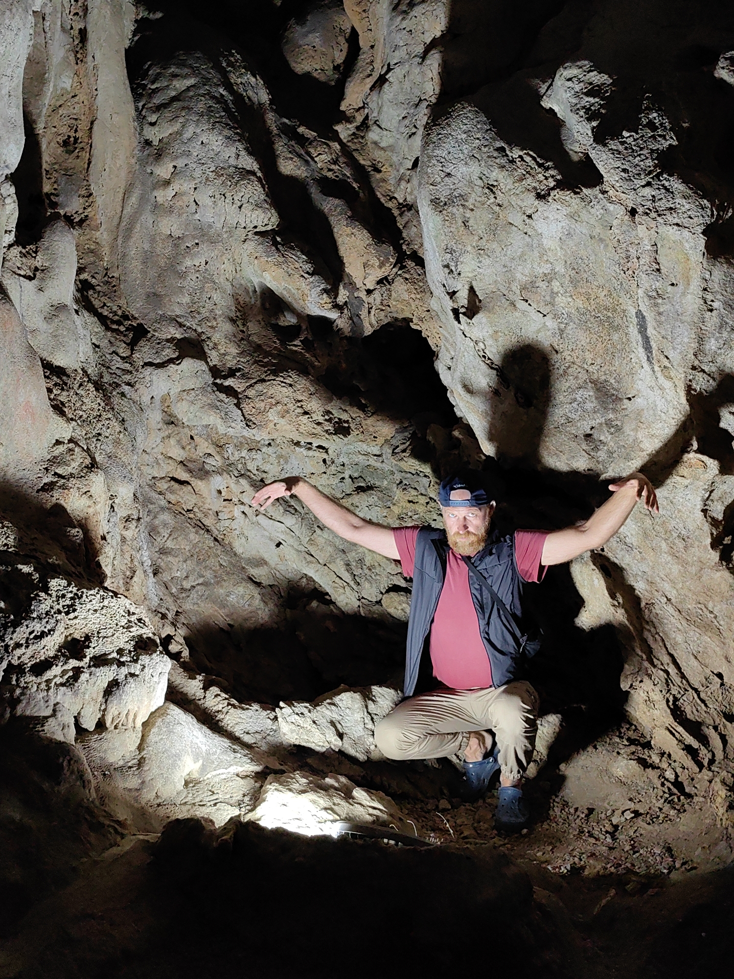 A man is crouching with arms outstretched in a dark cave in Lạng Sơn, Vietnam. The man has a beard and is wearing a baseball cap, a red shirt, a dark vest, and tan pants. He is illuminated by a light source that is located in front of him, and a light source to the upper right that is casting a shadow of his head and arms on the cave wall. The cave walls are rugged and appear to be made of rock.