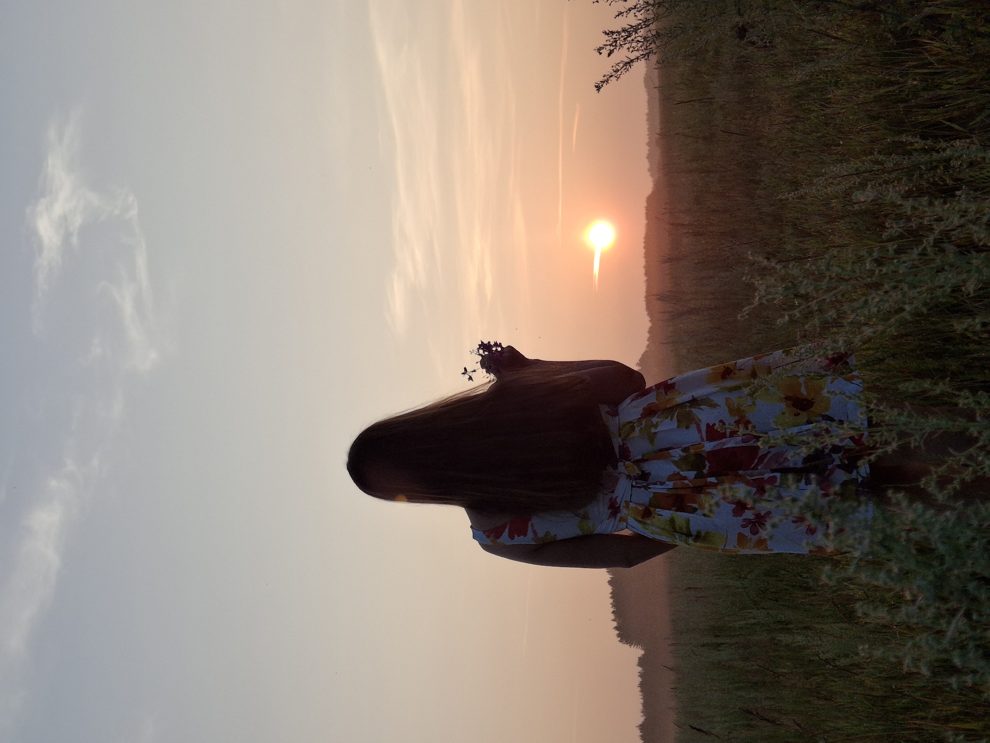The image depicts a young woman standing in a grassy field at sunset, facing away from the camera. She has long, dark hair and is wearing a floral print dress. She is holding a small bunch of wildflowers in her right hand. The sun is low on the horizon, casting a warm glow over the landscape. In the distance, a line of trees can be seen, shrouded in a light mist. The sky is a soft gradient of pink and orange, with a few wispy clouds. The overall impression is one of peace and tranquility. The location is identified as Plishkino, Russia.