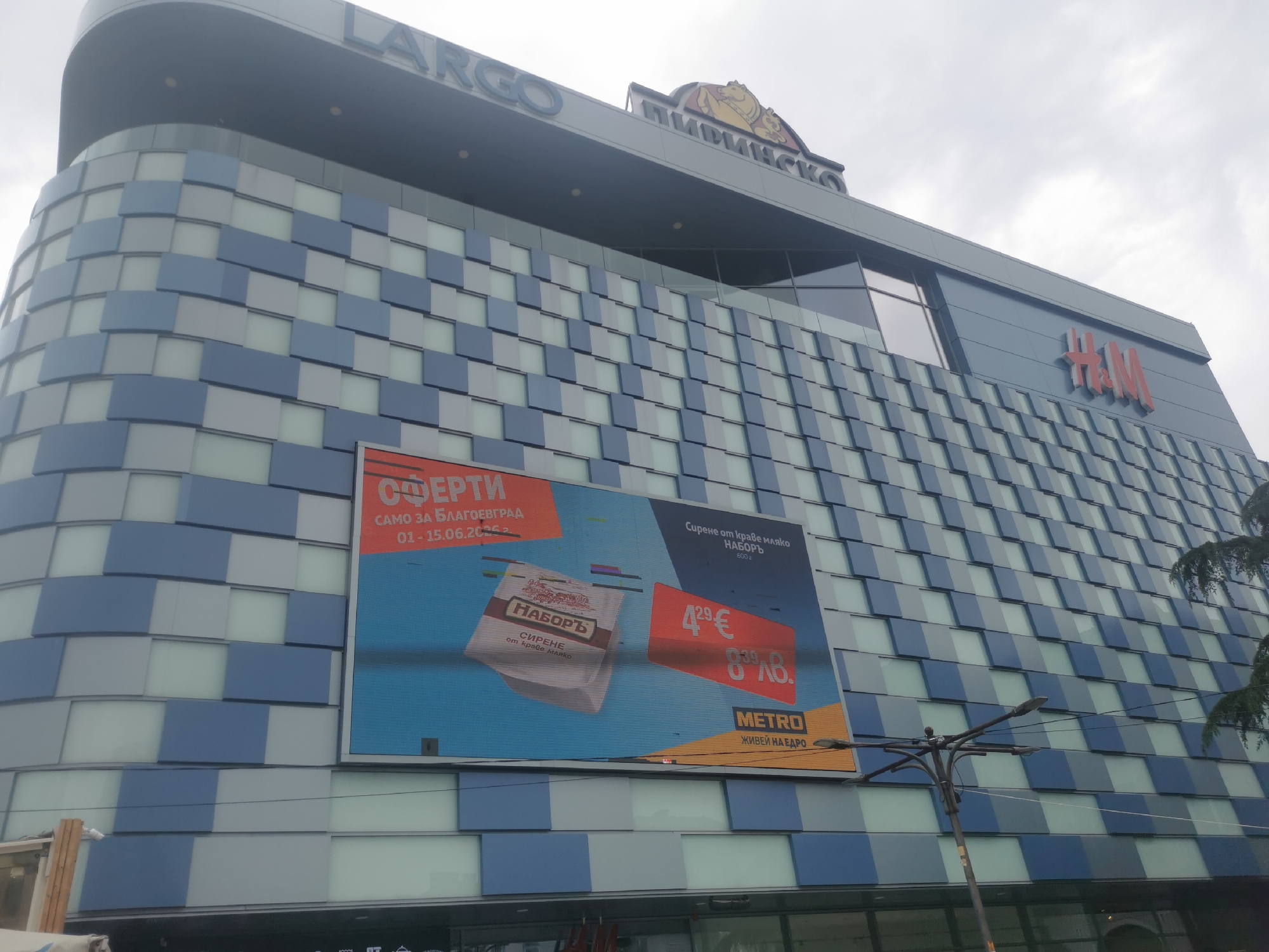 This image captures the exterior of a modern commercial building, likely a shopping mall, in Blagoevgrad, Bulgaria, under an overcast sky during daytime. The building features a distinctive facade composed of alternating blue, light blue, and light gray rectangular panels, giving it a patterned, geometric appearance.
Prominently displayed on the top of the building are several large signs. "LARGO" is visible on the upper left, while a partially obscured sign with a lion logo and Cyrillic text "ПИРИНСКО" (Pirin Beer) is in the center. To the upper right, the red "H&M" logo is clearly visible, indicating a retail store within.
A large digital billboard occupies a significant portion of the building's facade. It displays an advertisement featuring an offer for "НАБОРЪ" (Nabor) brand "Сирене от краве мляко" (cheese from cow's milk), specifically 600g. The advertisement shows two prices: "4.29€" (Euro) and "8.39лв." (Bulgarian Lev). Text on the billboard states "ОФЕРТИ" (Offers) and "САМО ЗА БЛАГОЕВГРАД" (Only for Blagoevgrad), along with a date range "01-15.06.2023". The advertisement is for "METRO" (Cash & Carry) and includes their slogan "ЖИВЕЙ НА ЕДРО" (Live wholesale).
In the foreground, a dark street light pole partially obstructs the lower right portion of the billboard. Some green foliage from a tree is visible on the far right. The overall scene depicts a functional urban commercial environment.