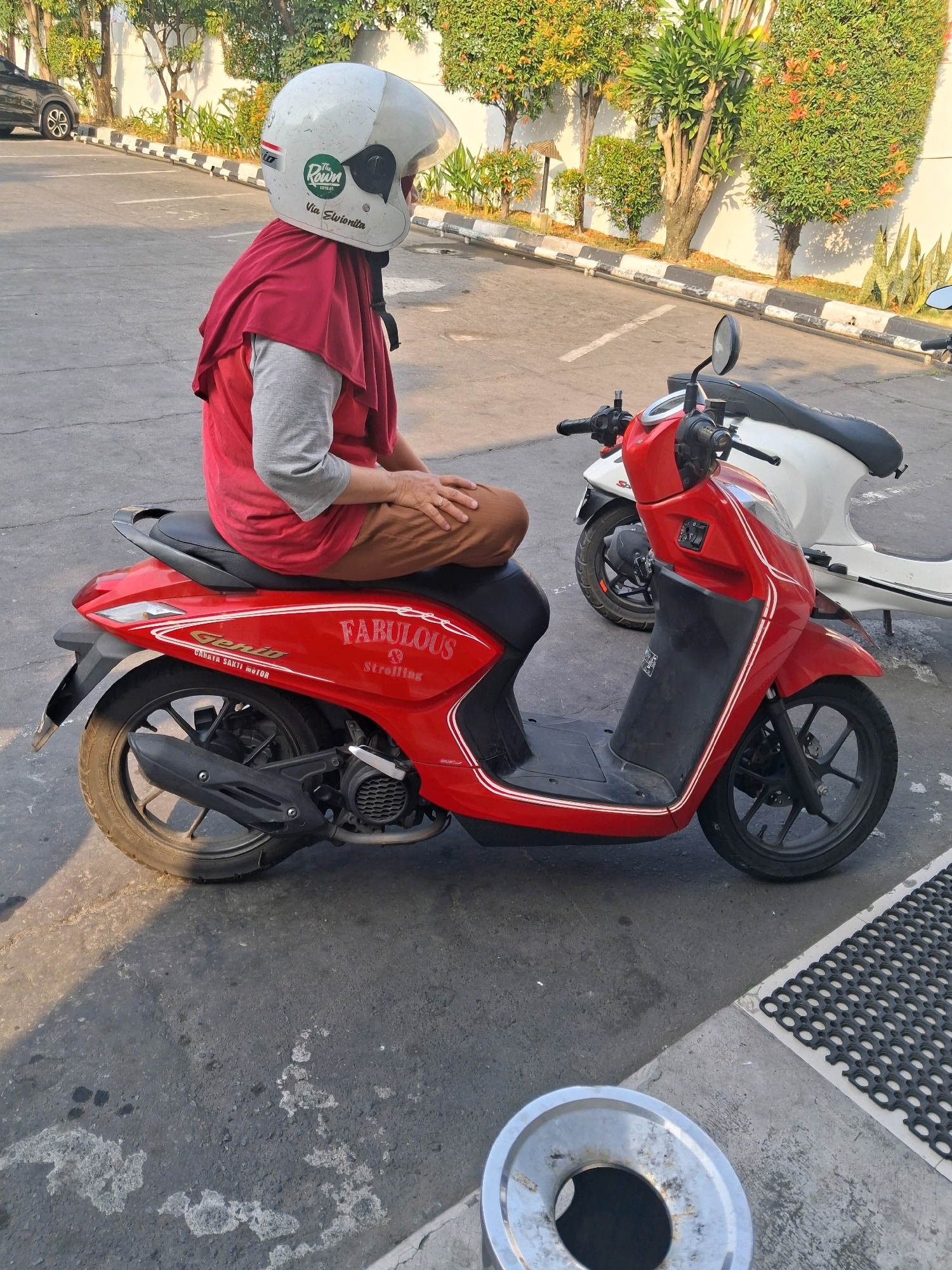 A person wearing a red headscarf and gray long-sleeved shirt, brown pants, and a white helmet with green and white stickers is sitting on a red scooter. The scooter has "Fabulous Strolling" and "Gento Cahaya Sakti Motor" written on its side. A white scooter is parked behind it. The scooters are on an asphalt surface in what appears to be a parking lot. In the background, there is a white wall, trees with orange and green foliage, and a black and white striped curb. The scene suggests a location in Surakarta, Indonesia, given the vegetation and style of the scooters. The lighting indicates it is daytime, with shadows cast on the ground.