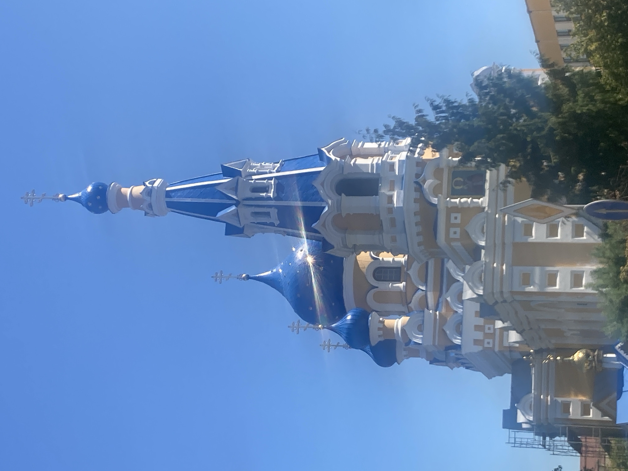 The image shows the Assumption Cathedral in Brest, Belarus. It is a large, ornate Orthodox church with multiple blue domes topped with crosses. The main tower is tall and pointed, also featuring blue and white accents. The building is predominantly yellow and white with decorative trim. A tall evergreen tree stands in the foreground, partially obscuring the right side of the cathedral. The sky is a clear, bright blue. In the lower left, scaffolding is visible on a section of the building, suggesting ongoing construction or maintenance. Traffic signs are also present in the foreground. The overall scene depicts a sunny day in a city.