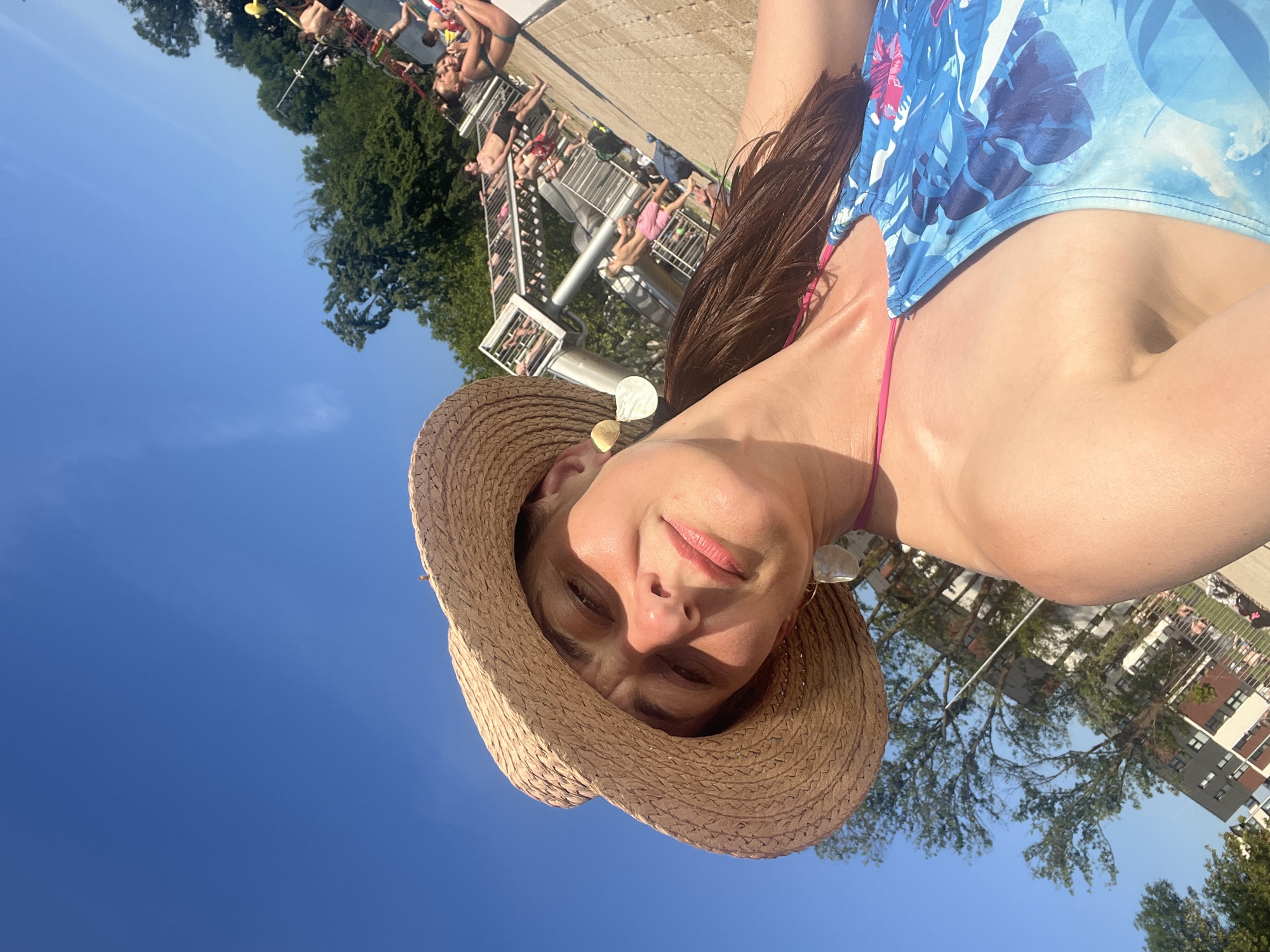 A woman wearing a straw hat and a blue floral swimsuit is taking a selfie outdoors on a sunny day. She is leaning back slightly, with the sky and trees in the background. Other people are visible in the background, some sunbathing and some near a water slide structure. The location is an outdoor public swimming pool.