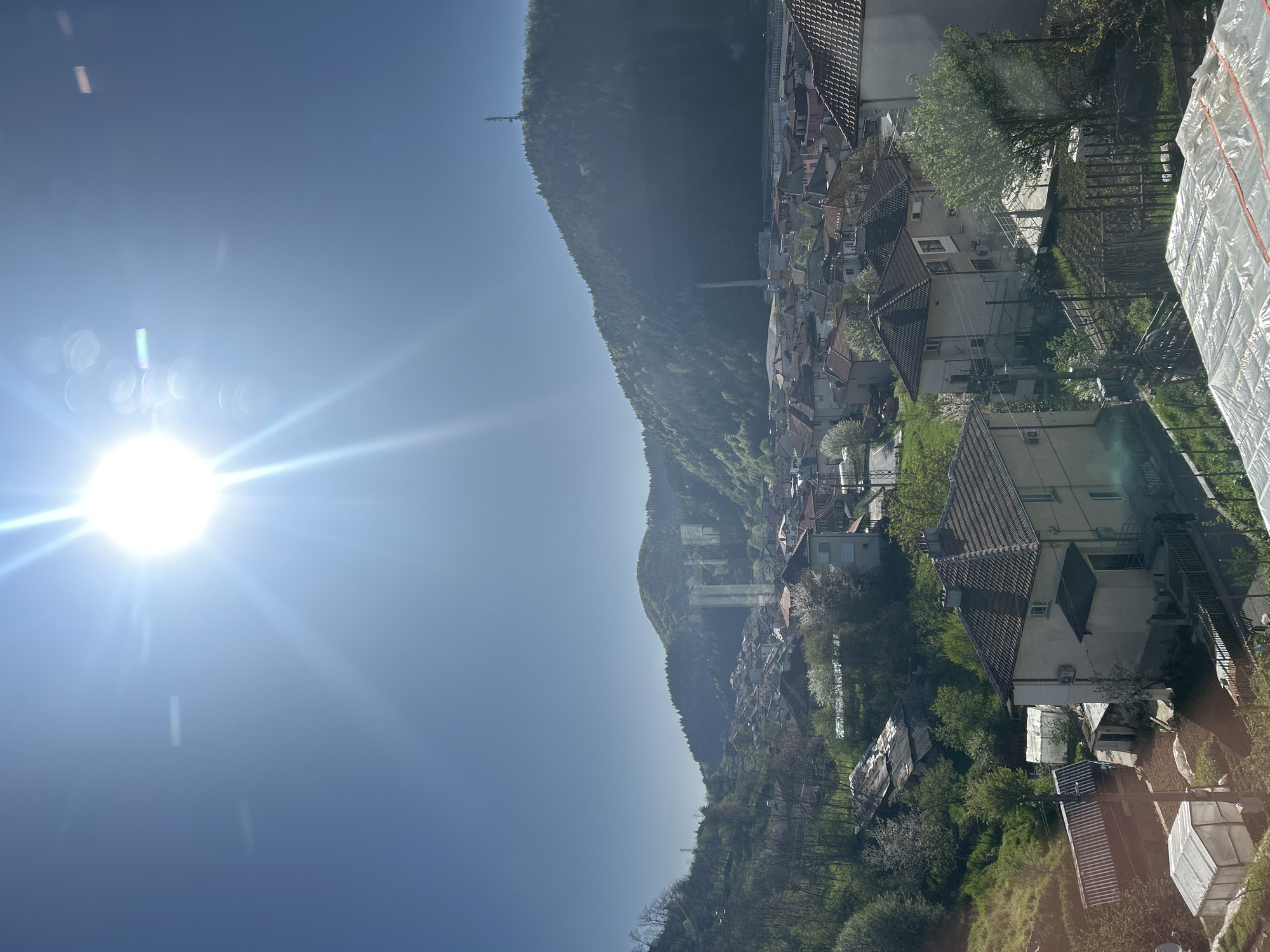 The image provides a wide view of Zlatograd, Bulgaria, a town nestled within a mountainous valley under a brilliant, clear blue sky. The sun, a powerful starburst of light with noticeable lens flare and light streaks, is prominently positioned in the upper left, suggesting bright daylight, likely mid-morning or early afternoon.
The town itself is characterized by numerous houses with distinctive reddish-brown tiled roofs and light-colored facades, densely spread across the valley floor and extending up the lower slopes of the surrounding mountains. Interspersed among the buildings is abundant green vegetation, including trees with fresh spring foliage, some showing white blossoms, indicating the season is spring. Power lines and utility poles are visible threading through the residential areas.
The backdrop consists of majestic, tree-covered mountains. A communication tower or similar structure is visible atop one of the more distant peaks. Faintly visible in the mid-distance is a bridge-like structure. In the immediate foreground on the right, there's a glimpse of what appears to be a yard or construction area, featuring white plastic sheeting covering materials, along with a metal fence and more greenery.
The scene is peaceful and serene, showcasing the picturesque blend of urban development and natural landscape. No specific human activity is depicted. There is no visible text in the image.