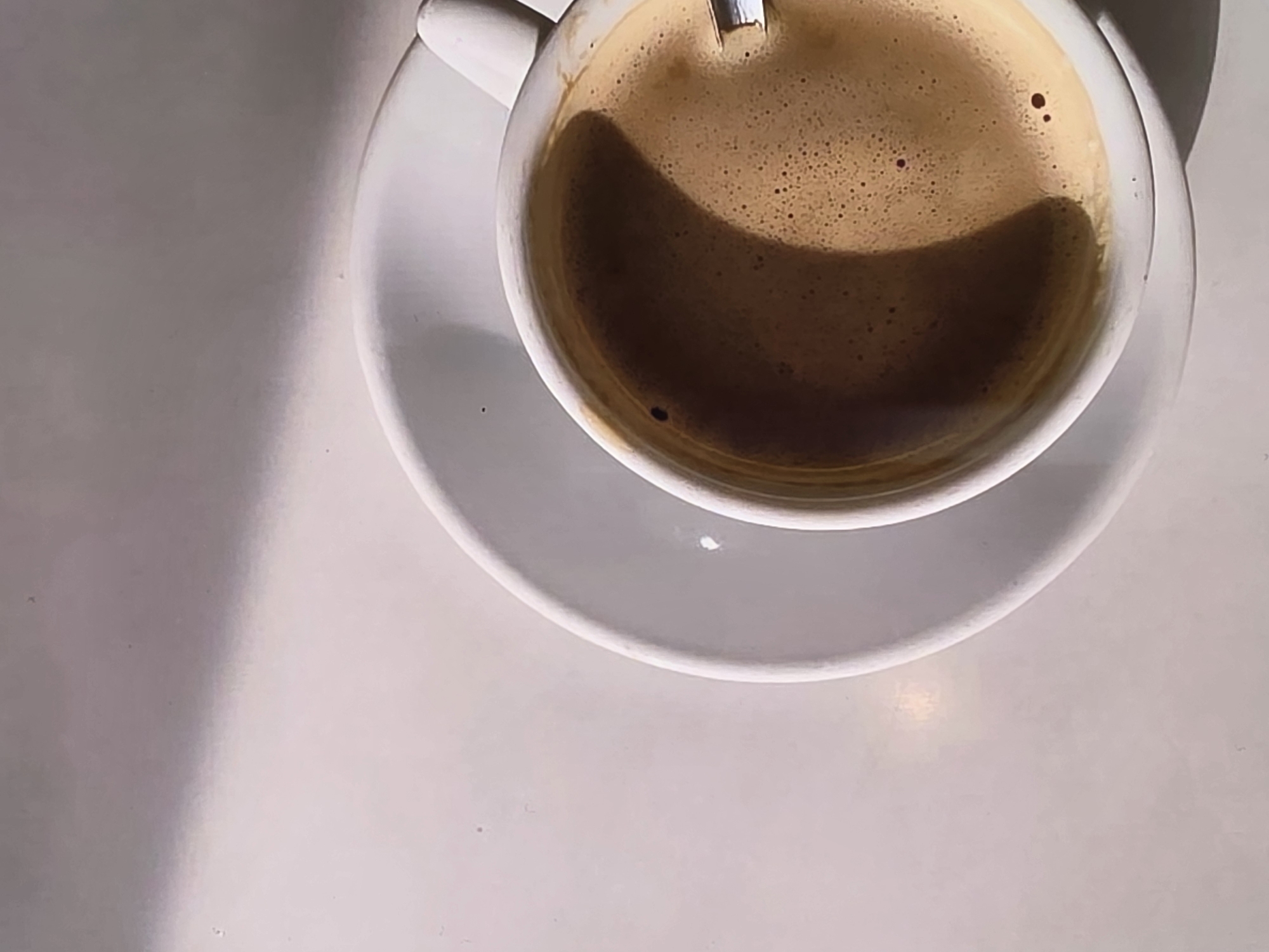 A close-up, overhead shot features a white ceramic cup of coffee sitting on a white saucer. The coffee has a frothy, dark brown crema and a small spoon is partially submerged in it. A shadow casts a diagonal line across the upper left portion of the image, creating a play of light and shadow on the white saucer and the background. The image does not contain any elements that would indicate its location in Voluntari, Romania, or any specific city.