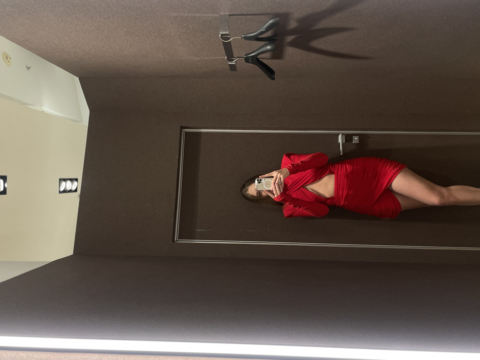 A mirror reflects a woman in a red dress, holding a phone to take a picture of herself. The reflection also captures a coat hanger and track lighting on a brown ceiling. A white wall with recessed lights is visible. The mirror's frame is thin and silver. The woman's legs are prominent in the reflection, suggesting a full-length mirror.