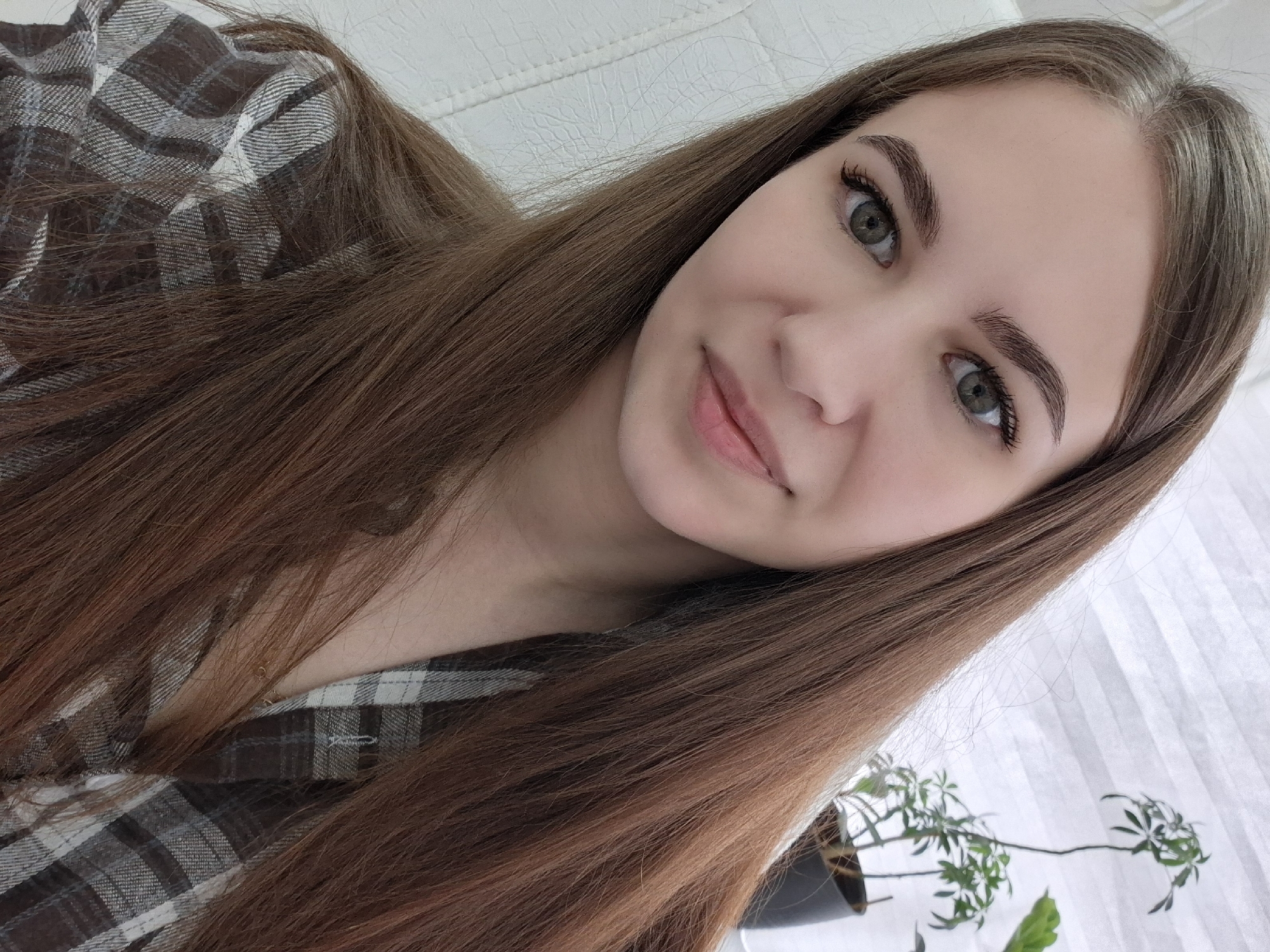 A young woman with long brown hair poses for a selfie. She is wearing a plaid shirt. Her skin is light, and she has light-colored eyes. A small potted plant is visible in the lower right corner of the frame. The location cannot be identified based on the image alone.