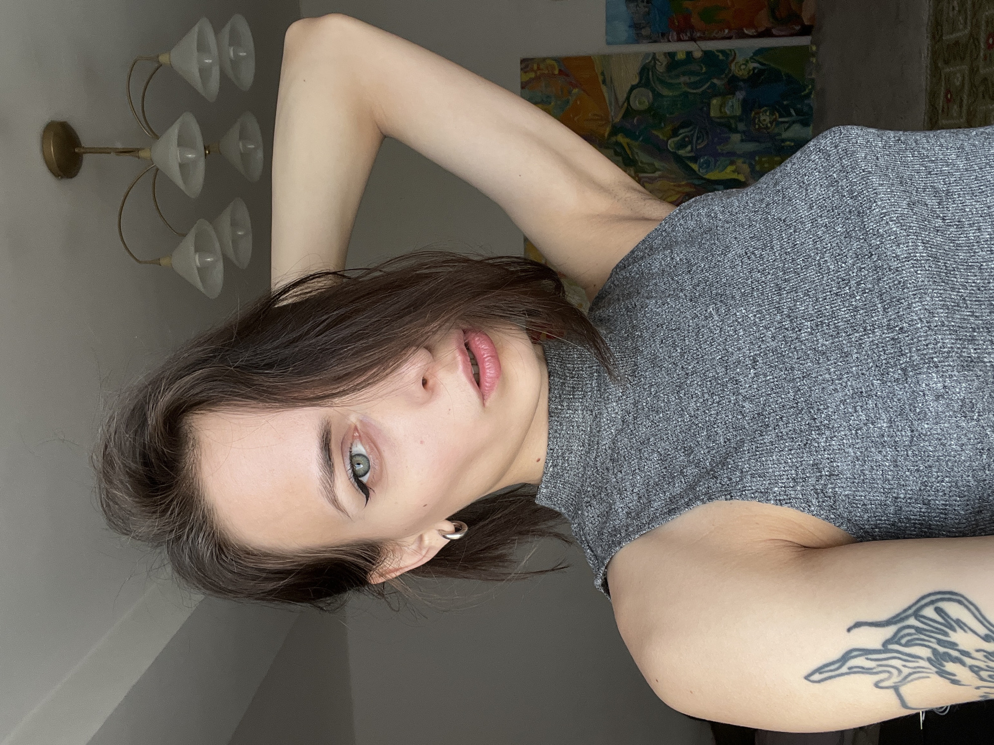 A person is positioned in front of a gray wall, wearing a gray turtleneck top. The person is angled to the left of the frame. A lighting fixture is visible on the left side of the image. An abstract artwork is partially visible in the background. The location is not provided in the image, so no description of Krasnoyarsk, Russia can be provided. A tattoo is visible on the arm of the person.