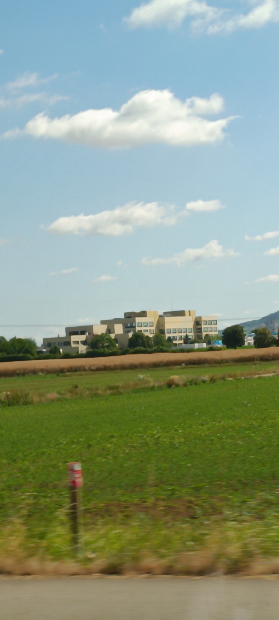 The image depicts a landscape scene with a large, modern building complex situated in the background. The building is light-colored, with multiple stories and numerous windows arranged in a grid pattern. It appears to be an institutional or commercial structure. In front of the building, there is a band of brown agricultural land, followed by a wide expanse of vibrant green grass that occupies the majority of the foreground. The grass is partially blurred, suggesting movement. A small, red and white striped marker is visible in the lower left portion of the grassy area. The sky is a clear blue with scattered white clouds. The overall impression is a rural or semi-rural setting with a significant man-made structure. It is not possible to determine the specific location within Fellbach, Germany from the image alone.