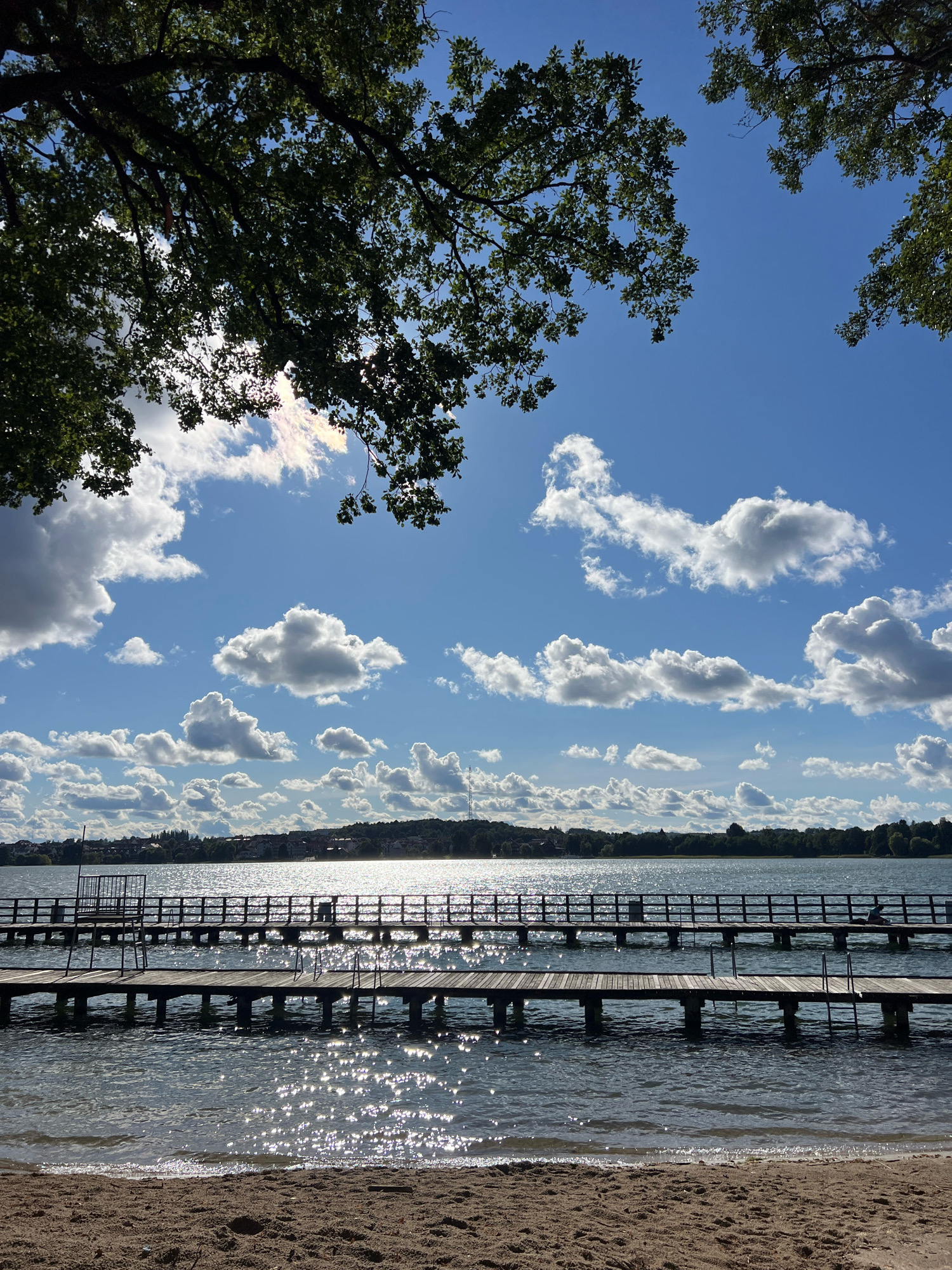 The image shows a wide view of a lake in Mrągowo, Poland, on a sunny day with scattered clouds. In the foreground, there is a sandy beach. Two wooden piers extend into the water, with their support structures visible beneath the surface. The lake's surface is shimmering with sunlight. In the middle ground, the distant shore is lined with trees and buildings. The sky is a vibrant blue, with fluffy white clouds. The top of the image is framed by dark green tree branches. There are a few people visible on the piers, appearing to be walking or sitting.