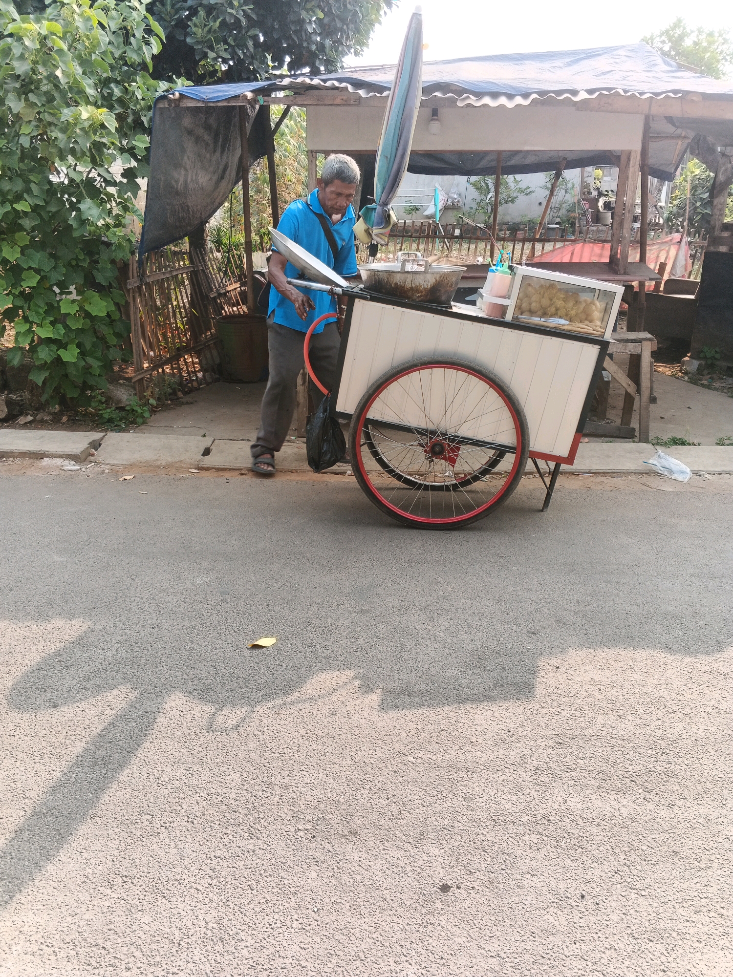 A man in a blue shirt and dark pants stands by a food cart on a paved road in Kadu, Indonesia. The food cart has a white body with red trim and a large spoked wheel with a red rim. It is topped with a canopy and a large, colorful umbrella. A metal basin is positioned on the cart, and a glass display case contains fried food items. Behind the cart, there is a makeshift stall with a blue tarp and bamboo fencing. Lush green foliage is visible to the left of the scene, and the ground is a mix of asphalt and concrete. Shadows are cast on the road from the right, indicating sunlight.