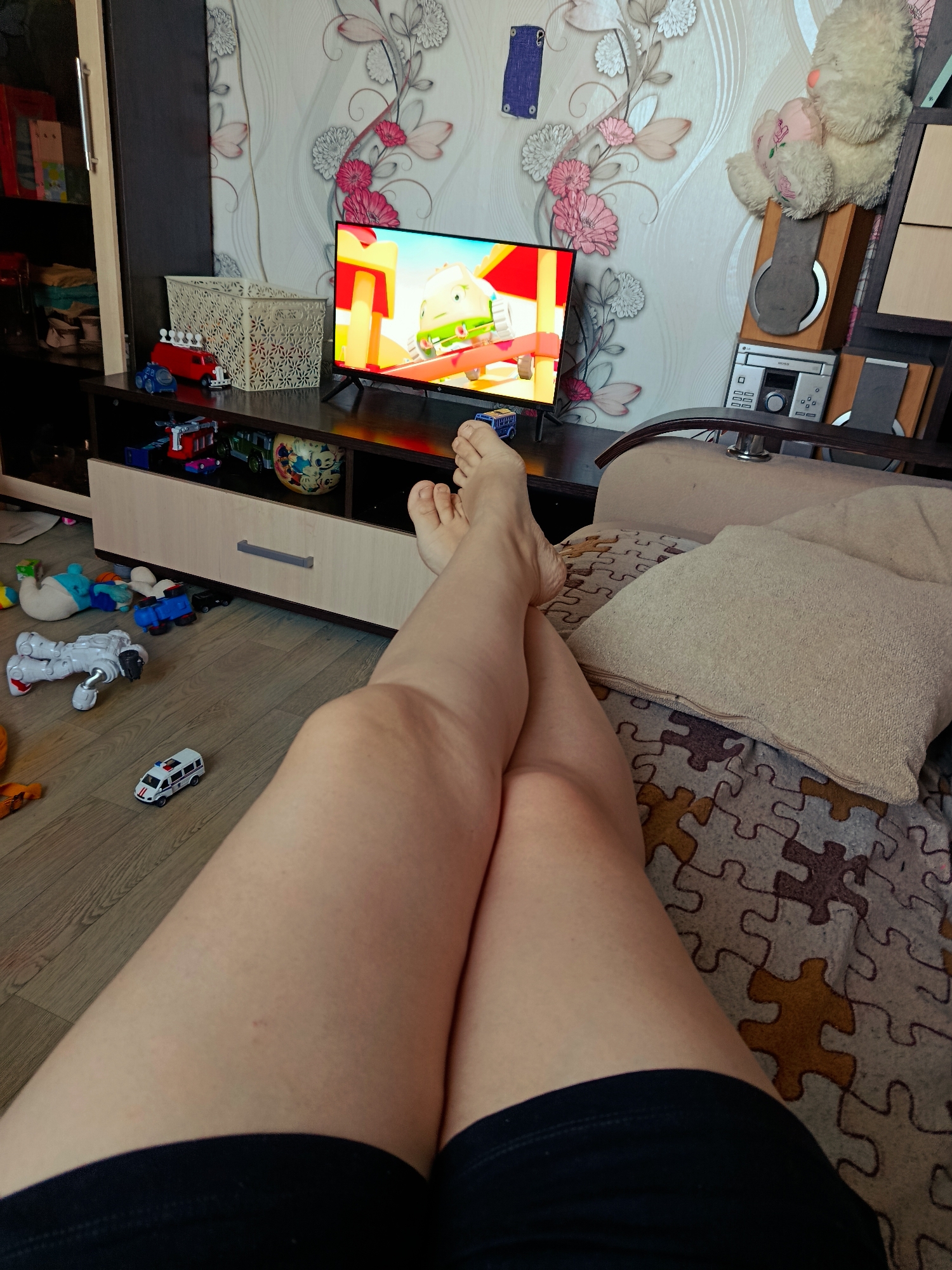 The image captures a first-person perspective of someone relaxing in what appears to be a living room or bedroom in Foki, Russia. The viewer's legs, clad in dark shorts, are crossed at the ankles and extended into the foreground, resting on a patterned blanket covering a couch or armchair.
In the middle ground, a flat-screen television displays a bright, animated children's cartoon featuring an anthropomorphic vehicle. The TV is situated on a dark brown media console with light-colored drawers and shelves. To the left of the console, a tall cabinet holds books. To the right, a stereo system with "LG" branding is visible on its main unit, next to which a beige teddy bear is perched on a shelf. The wall behind the TV is adorned with wallpaper featuring an elegant floral design of pink and white flowers and green stems.
The light-colored wooden floor in front of the console is scattered with numerous children's toys, including a white robot, various small cars and trucks (one white ambulance toy clearly shows "03" and a red cross symbol), and other colorful play items. A white decorative basket containing more toys sits on top of the TV console, and additional toy vehicles like a red fire truck and a green truck are visible within the console's open shelves.
The activity taking place is one of leisure and relaxation, with the person comfortably watching television, likely a program geared towards children given the on-screen content and the prevalence of toys in the room. The overall atmosphere conveys a casual and family-friendly home environment.
