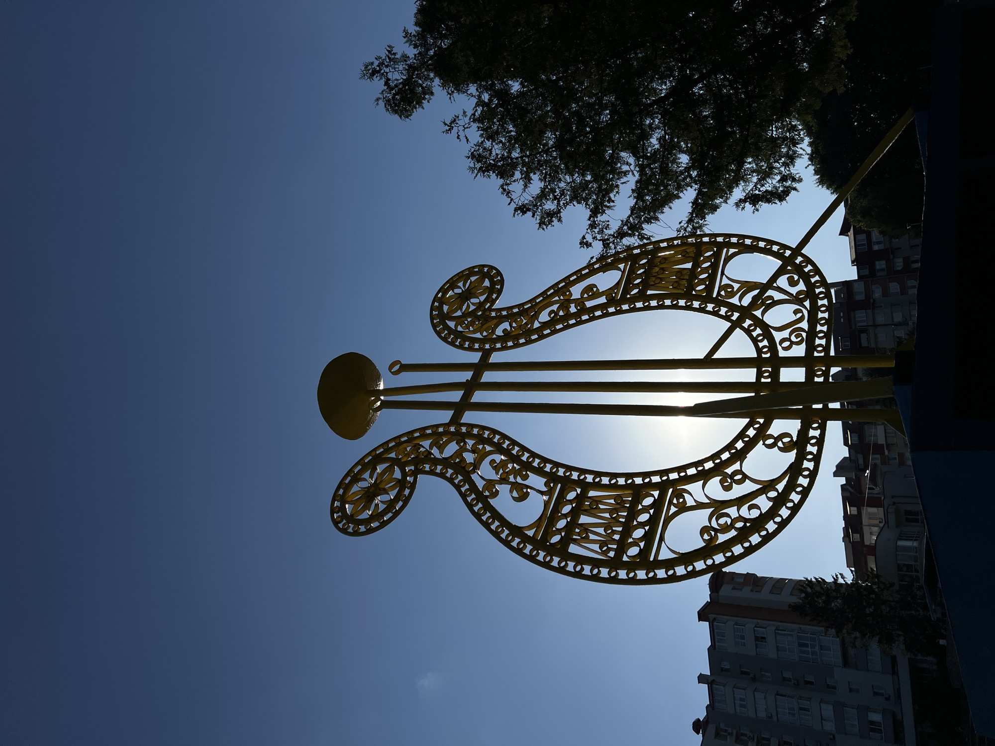 The image captures a vibrant yellow, lyre-shaped sculpture silhouetted against a bright blue sky. The sculpture, a prominent object, is intricately designed with decorative scrollwork and circular cutouts, suggesting an artistic and perhaps musical theme. Behind the sculpture, the branches of a dark green tree create a stark contrast against the clear sky, while the upper portions of several residential buildings are visible to the right and bottom of the frame. The sun is visible through the sculpture, creating a lens flare effect and indicating it is daytime, likely midday given the brightness. The clear sky suggests fair weather. The overall scene evokes a sense of public art installation in an urban setting. There is no visible text in the image. The context provided places this scene in Ternopil, Ukraine.