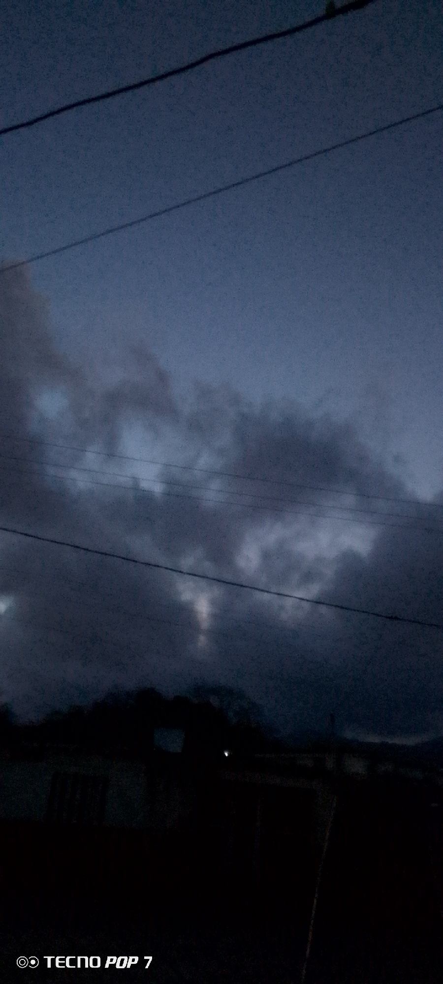 The image depicts a dusk or nighttime scene in what appears to be a residential area of Altagracia, Venezuela. The sky is dark with heavy, ominous clouds. Power lines are visible across the sky. In the foreground, there are dark silhouettes of buildings and vegetation, obscuring details. One faint light source is visible, likely from a distant building or street lamp. The overall impression is one of darkness and potential inclement weather. The "Tecno Pop 7" watermark suggests the photo was taken with a Tecno Pop 7 mobile device.