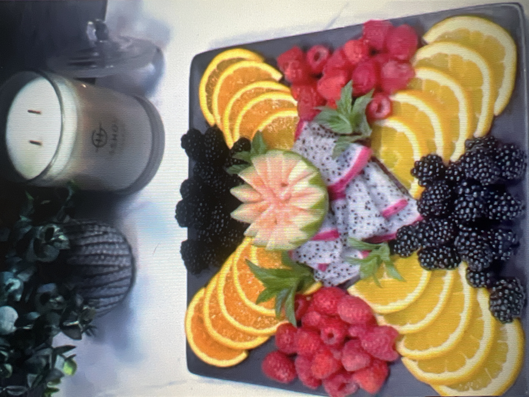 A fruit platter arranged in a butterfly shape is displayed on a dark rectangular plate. The platter includes sliced oranges arranged in a semi-circle, blackberries clustered at the top and bottom, raspberries scattered around the sides, and pieces of dragon fruit in the center. A decorative element, possibly a carved grapefruit, sits atop the dragon fruit, adorned with mint sprigs. The platter is placed on a light-colored surface, possibly a table, with a lit candle in a glass jar and a potted plant with dark green foliage visible in the background, suggesting an indoor setting. The overall scene appears to be a decorative food display.