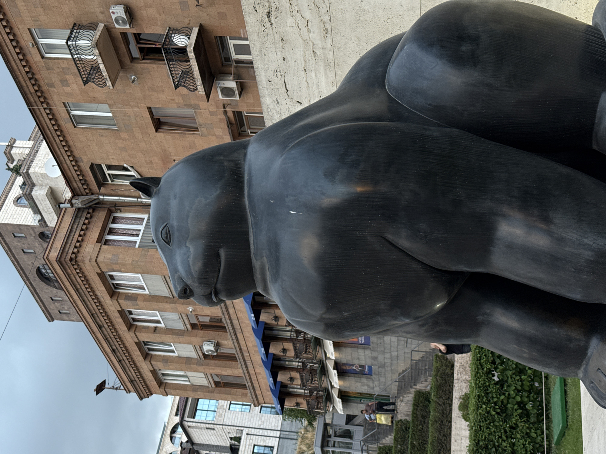 A large, dark-colored statue of a stylized animal, possibly a bear or panda, dominates the foreground, viewed from its left side and rear. The statue, made of a smooth, dark material like bronze, is depicted in a crouching or resting pose, with a gentle expression on its visible face.
In the mid-ground and background, several multi-story buildings with light brown or beige stone facades rise under an overcast sky. These buildings feature numerous windows, many with white frames, and ornate dark iron balconies. Multiple white air conditioning units are mounted on their exteriors. Below one of the buildings, blue awnings suggest storefronts, and a satellite dish is visible on a building further back.
Below the buildings and behind the statue, a public space is visible, characterized by light-colored stone paving, steps, and neatly trimmed green hedges. Two people are discernible in the lower right portion of the image, near these steps and greenery, with one person standing and another possibly sitting. The scene depicts a typical daytime setting in a city, consistent with the provided context of Yerevan, Armenia. No clearly legible text is discernible on any signs or banners.