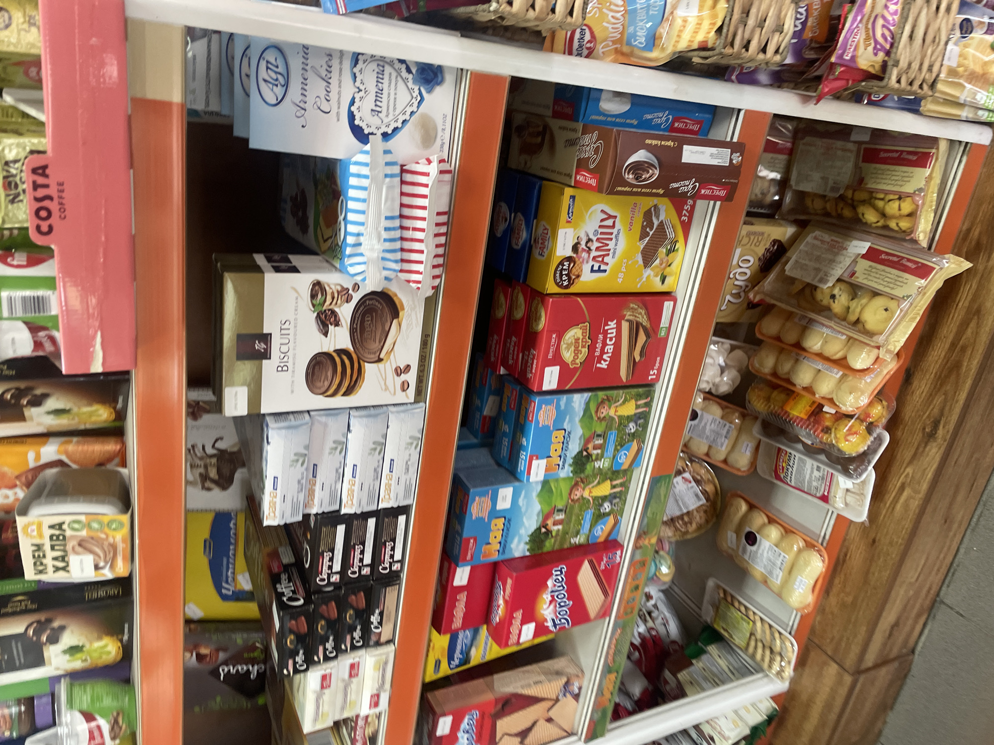 The image depicts a retail store aisle in Konstantinovo, Bulgaria, showcasing a variety of packaged food products, primarily sweets, cookies, wafers, and baked goods, arranged on multiple shelves and a lower display unit.
On the partially visible top shelf, products include a container labeled "КРЕМ ХАЛВА" (Cream Halva) and a pink shelf-edge sign for "COSTA COFFEE."
The second shelf, from left to right, displays several brands of biscuits and chocolates. A gold box is labeled "BISCUITS" featuring images of chocolate biscuits and a drink. To its right, multiple black boxes branded "Coffee" contain chocolates or small confectionery items. White boxes labeled "Трапеза" (Trapeza) are stacked on the far right. In the center, between the "BISCUITS" and "Трапеза" products, are two stacks of packaged cookies or wafers: one with red and white stripes and another with blue and white stripes. A blue box labeled "Agi Armenia Cookies" with a heart design and blue rose is visible on the far right.
The third shelf features various boxes of wafers and mini waffles. From left to right, a red box is labeled "Боровец" (Borovets), and another red box is labeled "Поген Край" (Pogen Kray), both displaying waffle images. A yellow box is branded "FAMILY mini waffles vanilla" with an image of wafers and cream. Adjacent to it are two blue boxes with cartoon graphics of children and houses, labeled "Паа" (Paa). A brown box with "ПРЕСТИЖ" (Prestigious) and a coffee cup graphic is also present.
The bottom display area consists of a wooden surface supporting various clear plastic containers and bags of individually wrapped baked goods, such as buns, small cakes, and possibly lokum, with labels visible but mostly unreadable. One label partially visible states "ЛУКУМ" (Lokum). To the far right, wicker baskets contain additional packaged items, including a "Dr. Oetker Pudding" mix. No individuals are visible in the scene. The shelving structure features orange shelf edges and white shelf surfaces.
